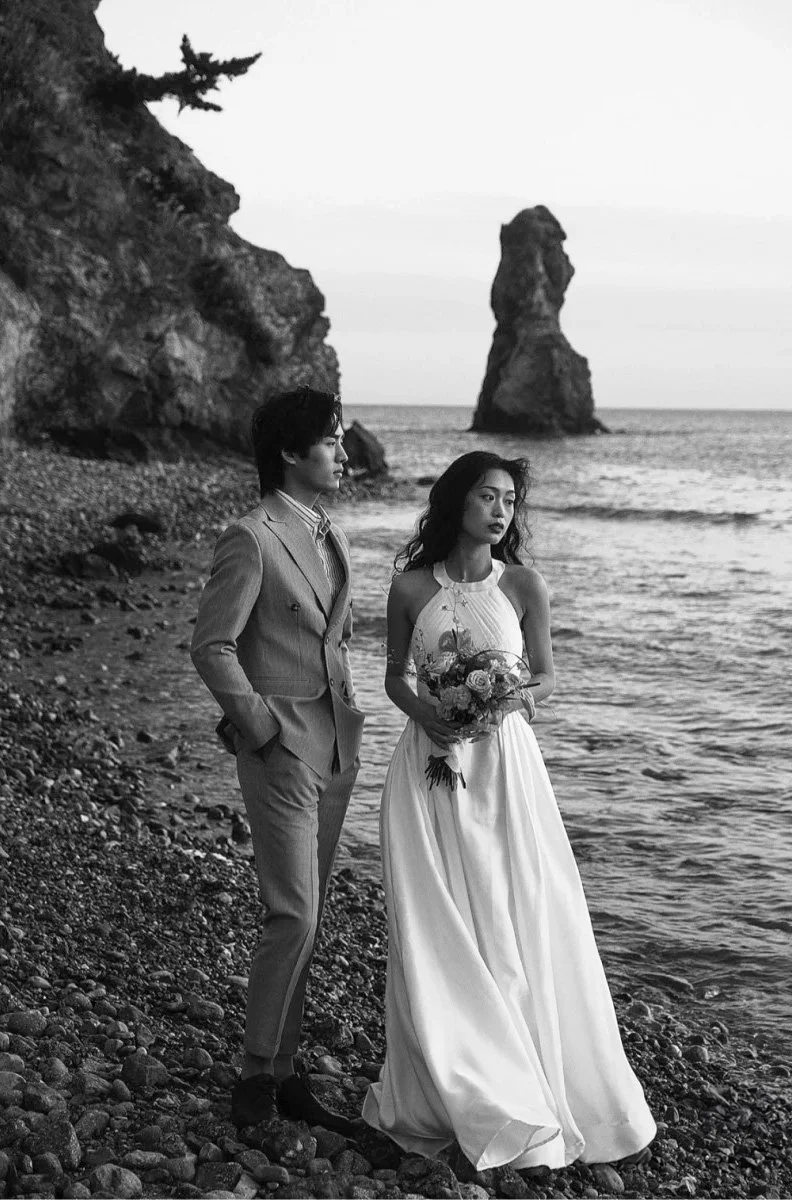 Black and white photo of a couple in wedding attire standing on a rocky beach, with a large rock formation in the sea and cliffs in the background.