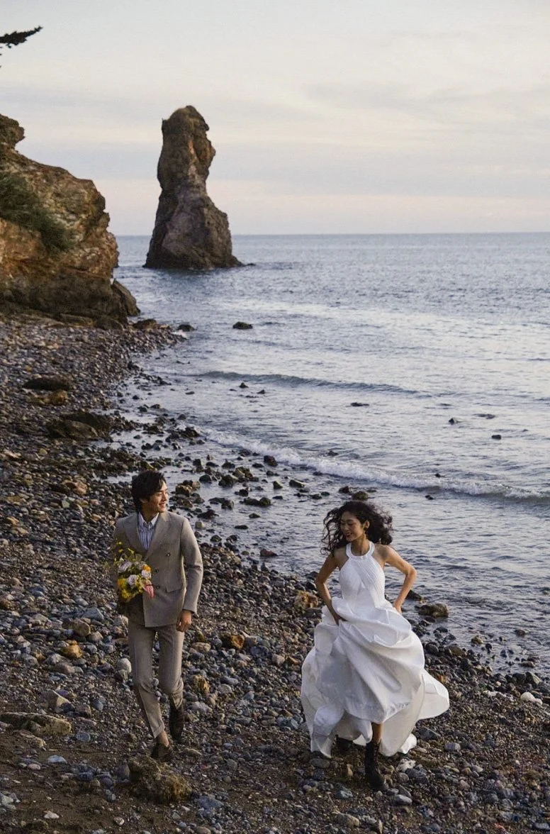 A man in a gray suit and a woman in a white wedding dress walk along a rocky beach with ocean waves, a large sea stack, and cliffs in the background.