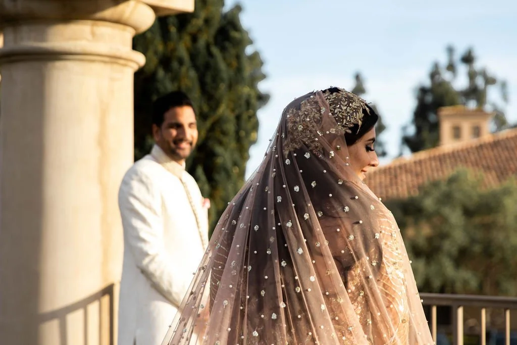 A bride wearing a sheer, embellished dupatta and a traditional outfit, smiling with a groom in a white sherwani in the background, outdoors with trees and a building in the distance.
