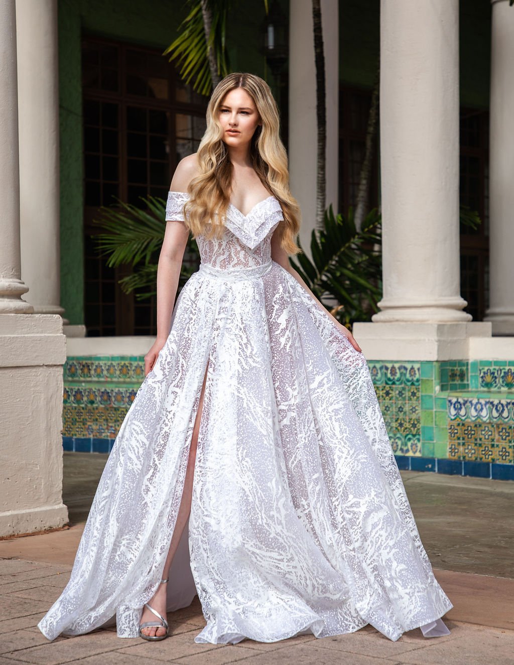 A woman with long blonde hair wearing a white lace off-shoulder gown with a slit, standing outdoors in front of columns and greenery.