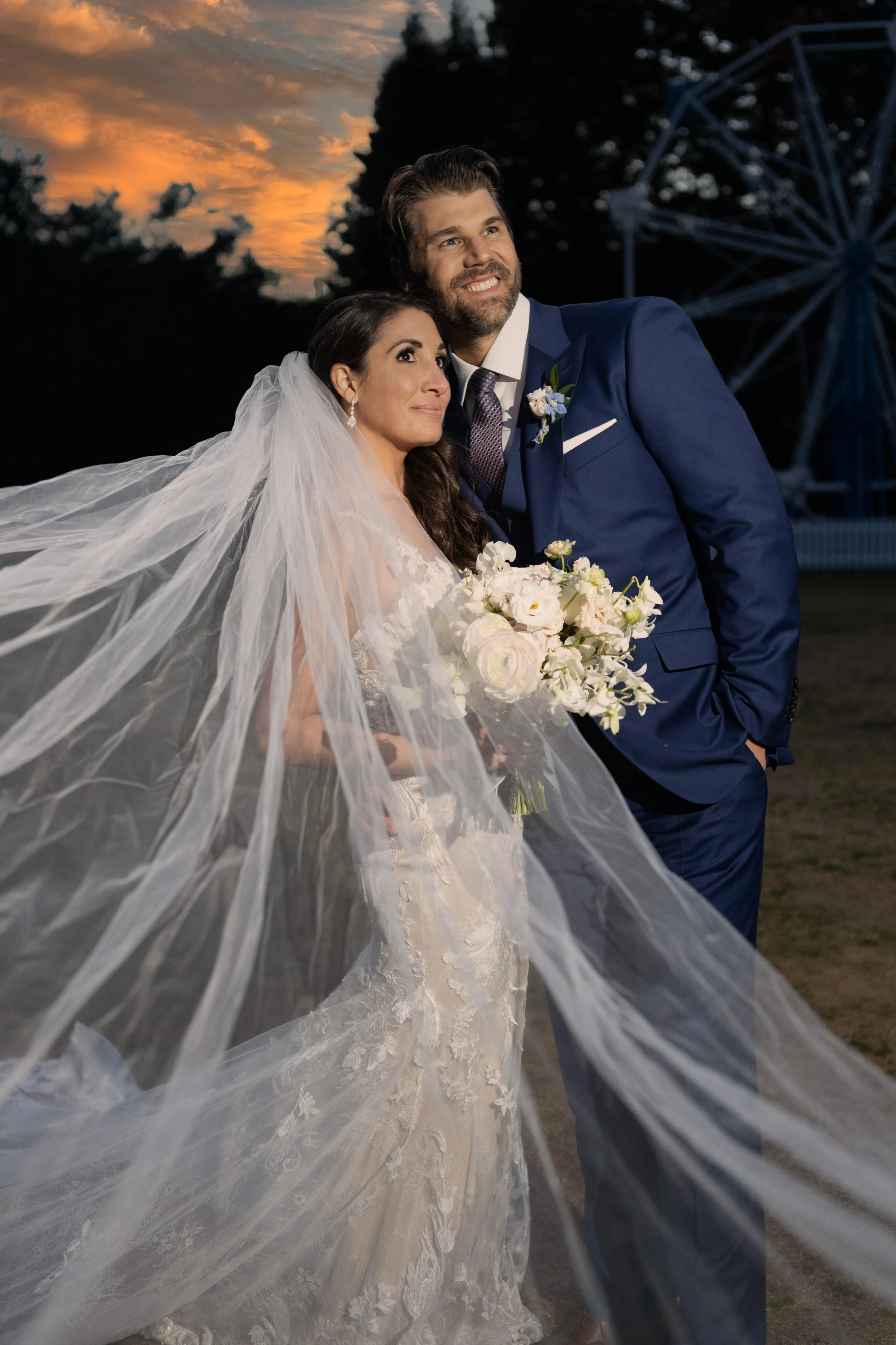 A newlywed couple posing outdoors at sunset, with the bride in a lace wedding gown and veil holding a bouquet of white flowers, and the groom in a blue suit smiling beside her, with a Ferris wheel in the background.