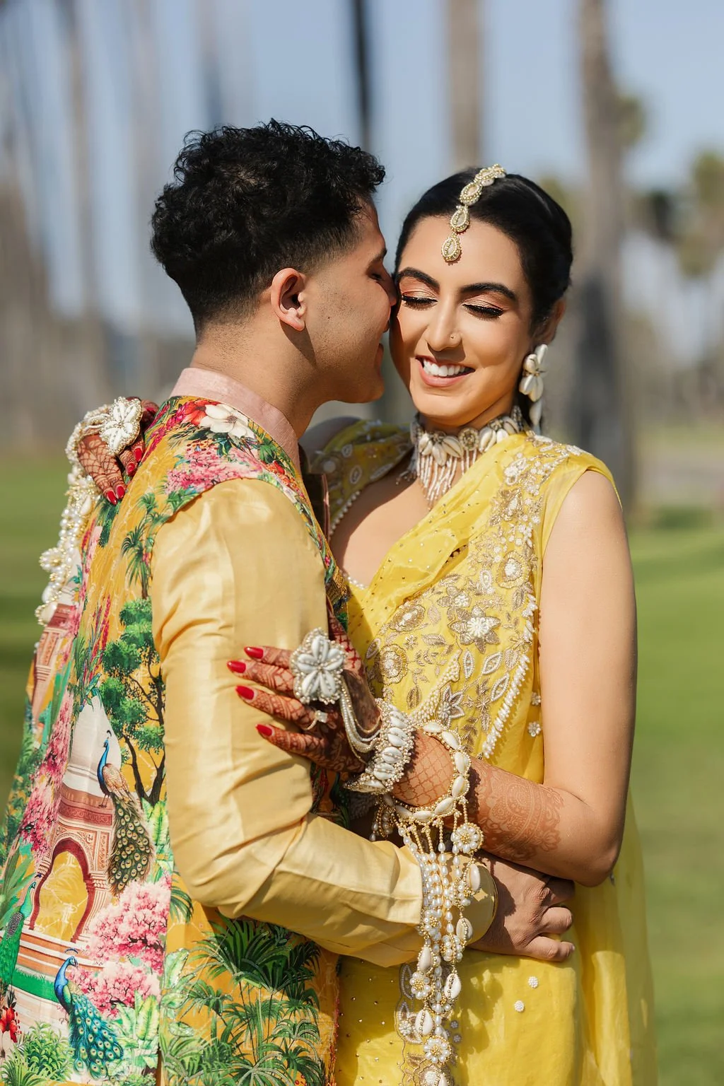 A couple dressed in colorful yellow traditional attire at their beach Haldi, sharing an intimate moment outdoors, smiling and embracing, with a blurred background of greenery and structures at Hilton Santa Barbara beachfront.