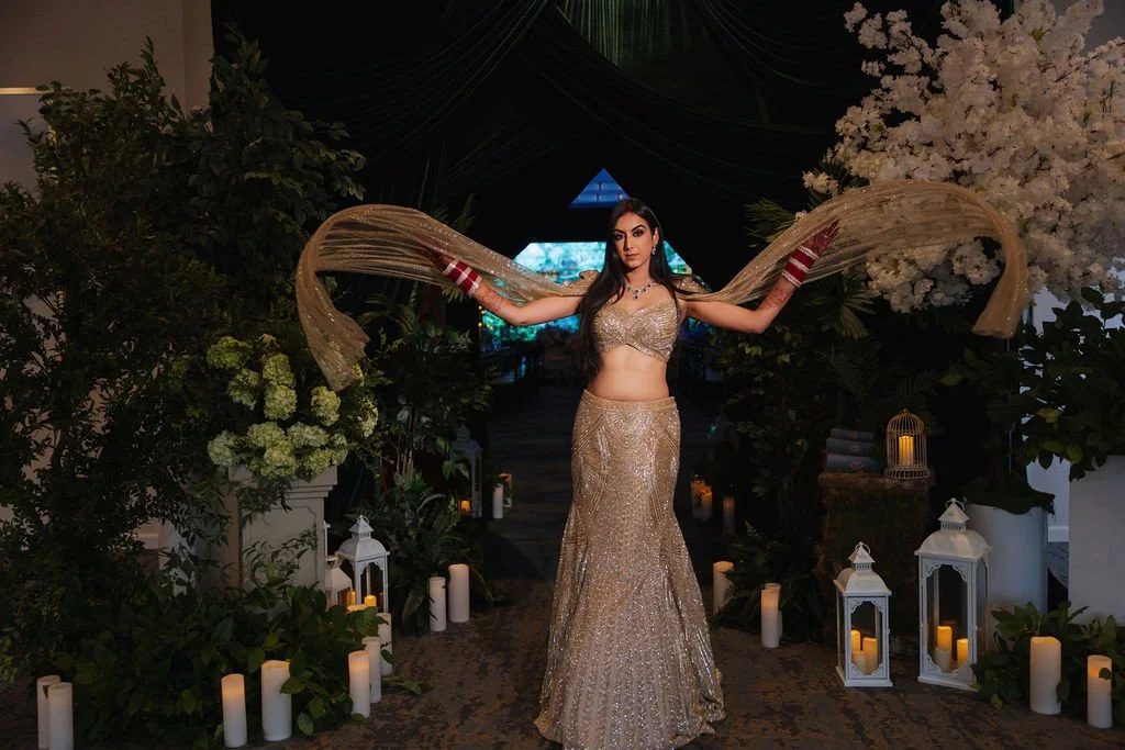 A woman in an ornate gold and silver traditional Indian outfit, with long flowing hair, stands in a decorated indoor setting with white lanterns and candles, surrounded by greenery and white flowers, holding her arms out to display her attire.