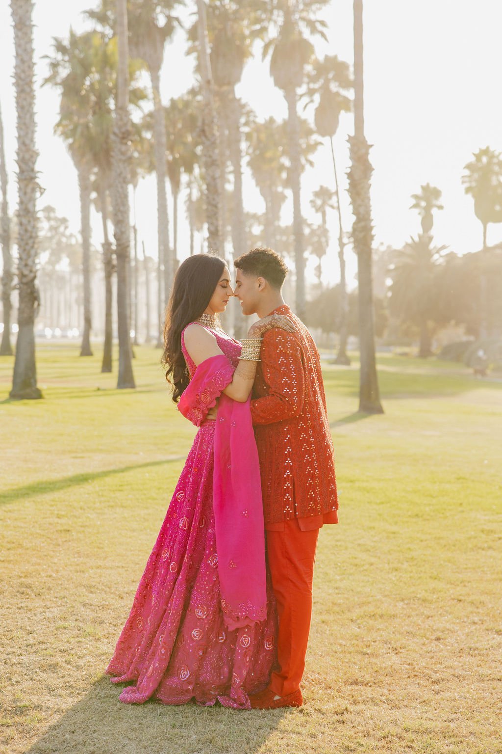 A couple dressed in colorful traditional Indian attire stands closely together outdoors on a grassy lawn with tall palm trees in the background during the golden hour of sunlight in Hilton Santa Barbara beachfront.