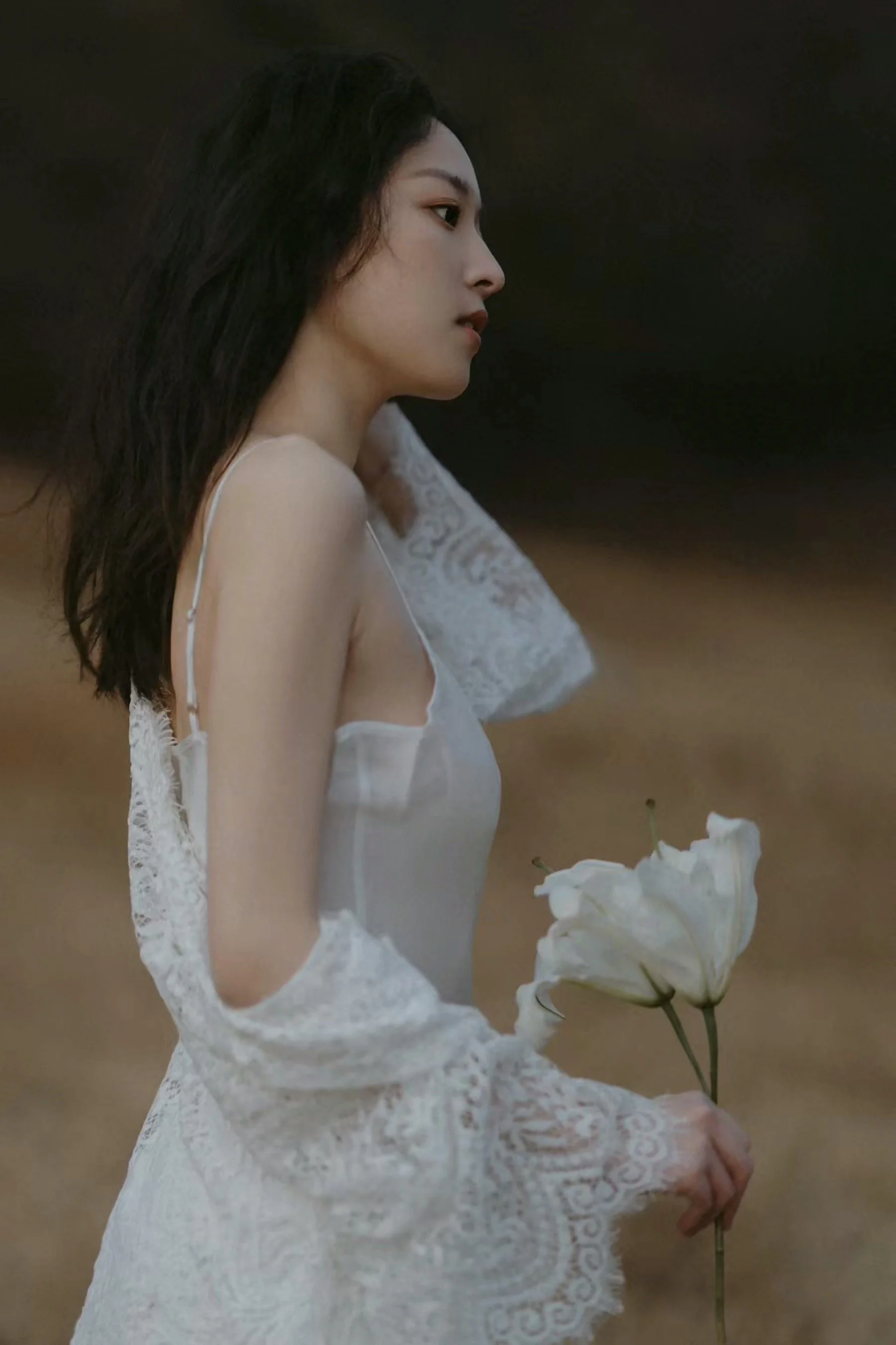 A woman in a white dress with lace sleeves holding white lilies, standing outdoors with a blurred natural background.