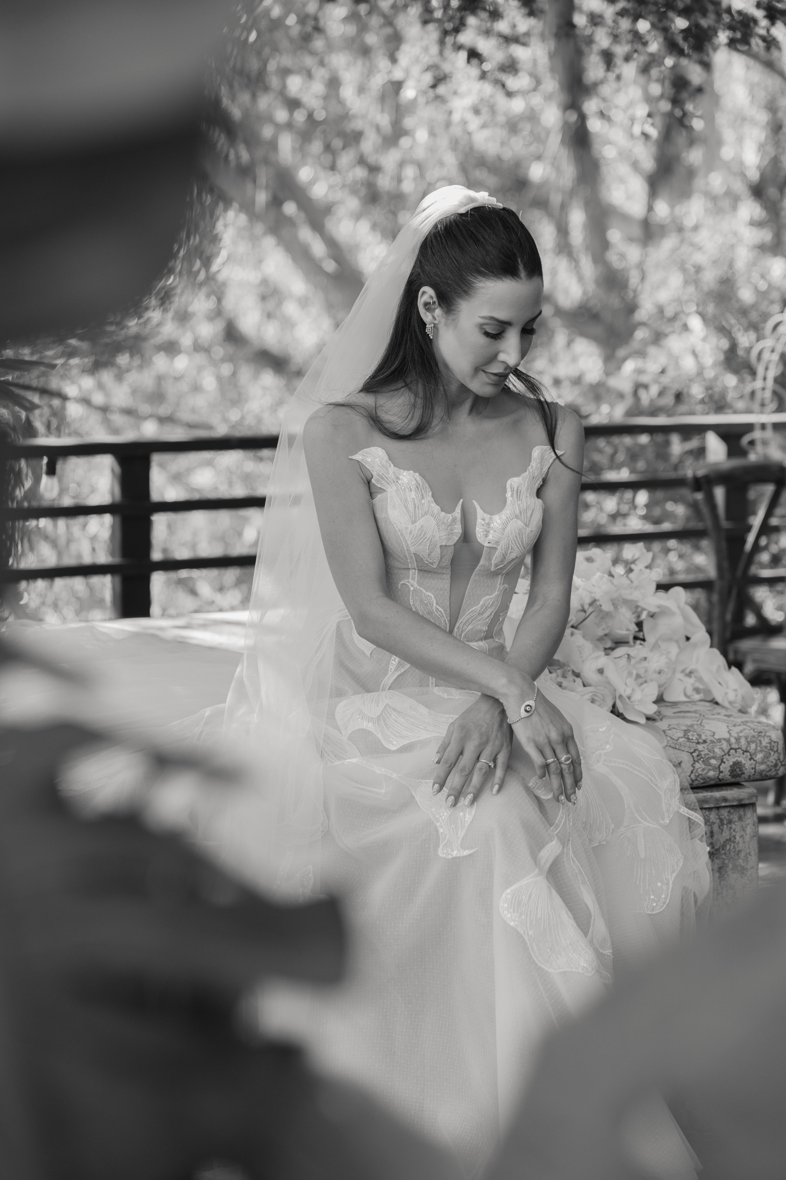 Black and white photo of a bride with dark hair in a wedding dress, sitting outdoors with trees in the background, looking down with her hands crossed in her lap.