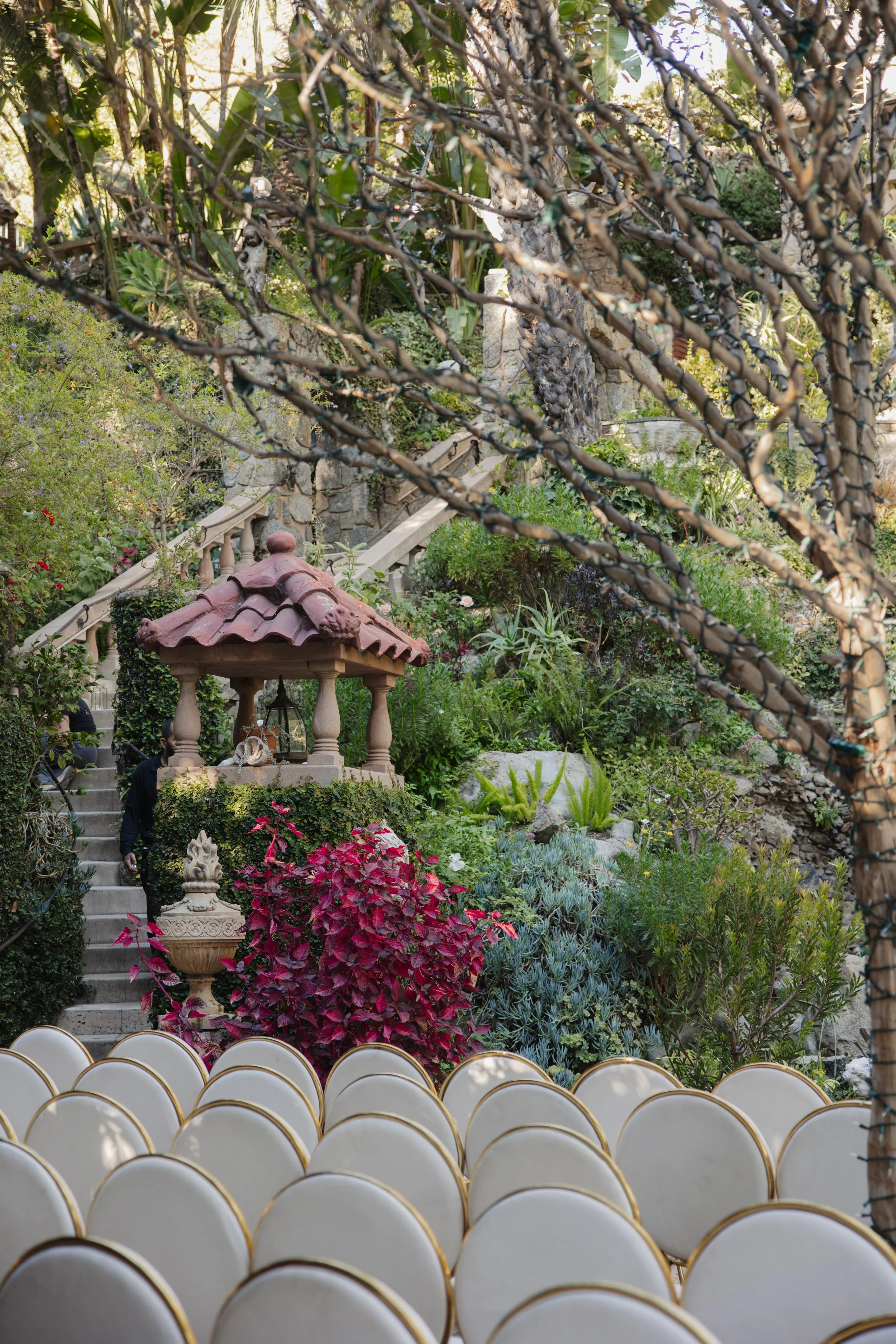 A lush garden with a terracotta tiled gazebo, stone stairs, green and purple plants, and white chairs with gold trim in the foreground.