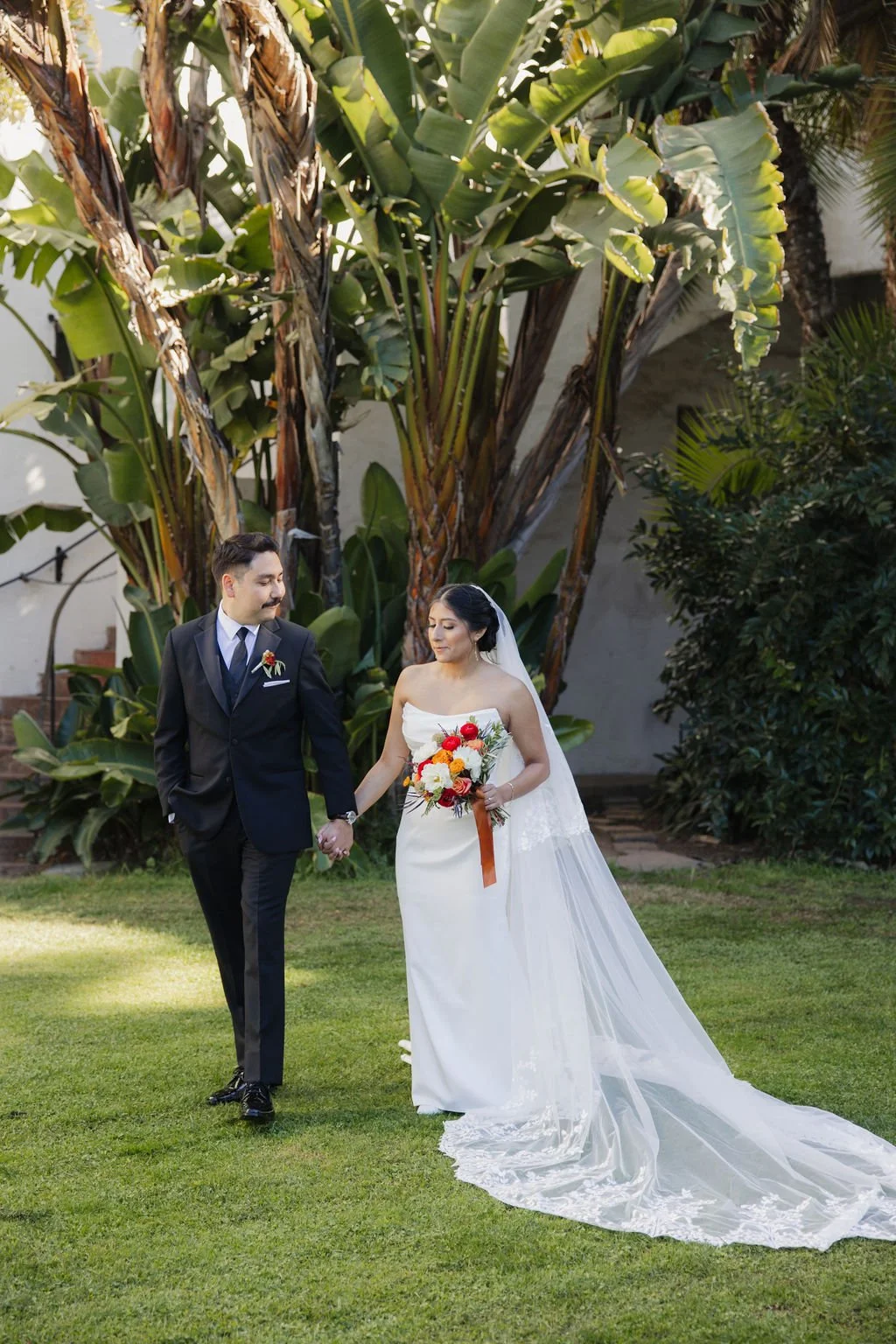A bride and groom holding hands walking on grass in a lush garden, with tropical plants and trees in the background.