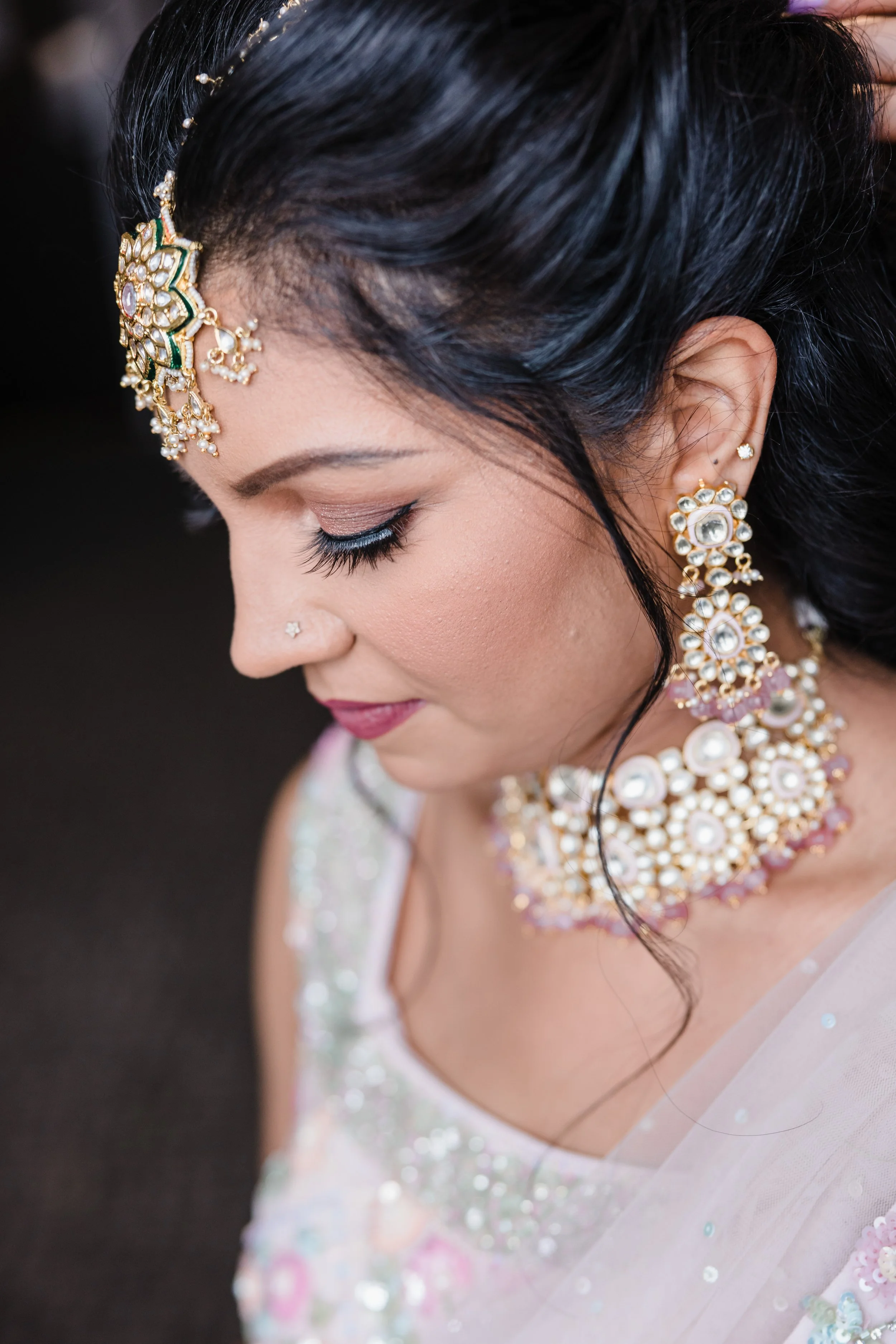 Close-up of a woman with dark hair wearing intricate traditional jewelry, including earrings, necklace, and headpiece, with her eyes closed and a subtle smile.