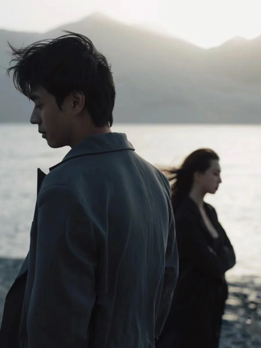 A young man and woman stand apart near a body of water, with mountains in the background. The young man has dark, disheveled hair and is wearing a jacket, looking down with a somber expression. The woman has long hair and is also dressed in dark clot