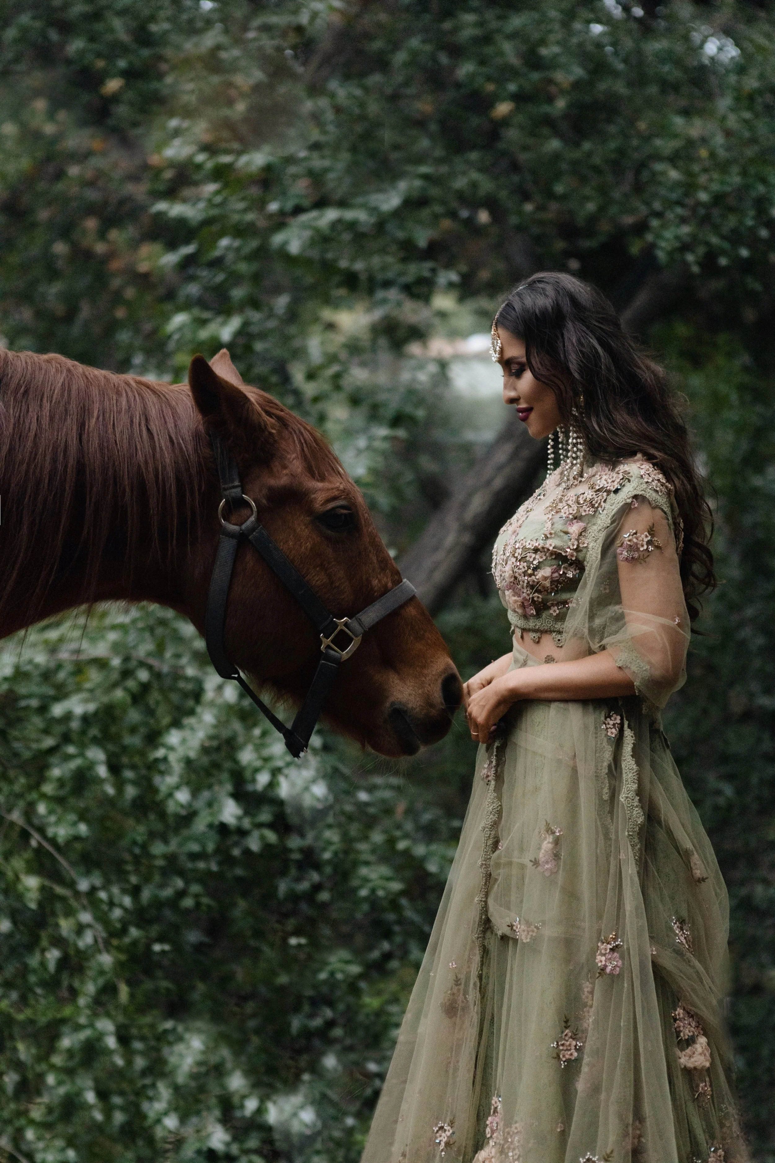 A woman in a long, embroidered green dress and jewelry stands in a forest, gently touching a brown horse.