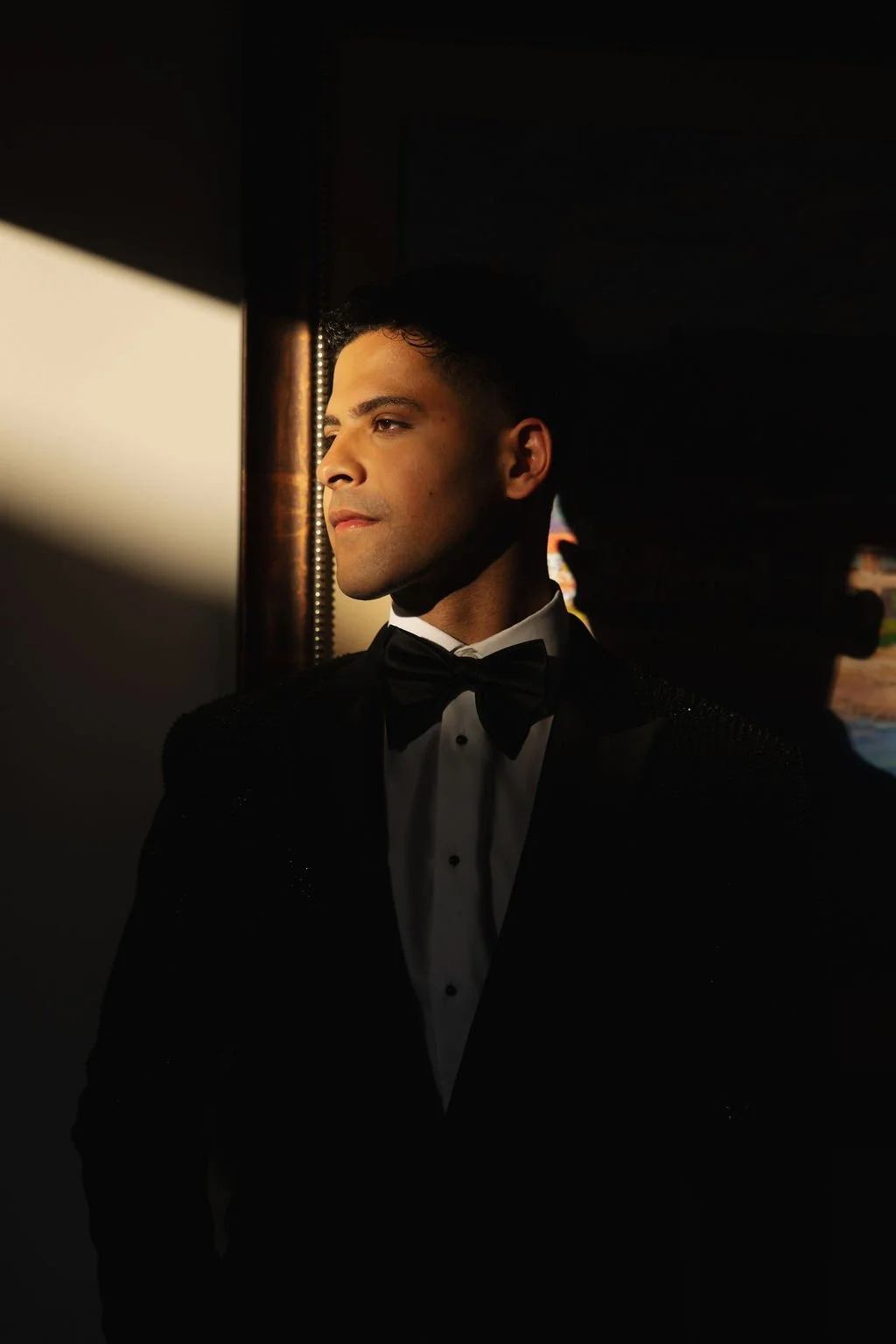 a chic Groom in tuxedo and bow tie looking out of a window at his wedding reception, with sunlight illuminating his face at Hilton Santa Barbara beachfront.