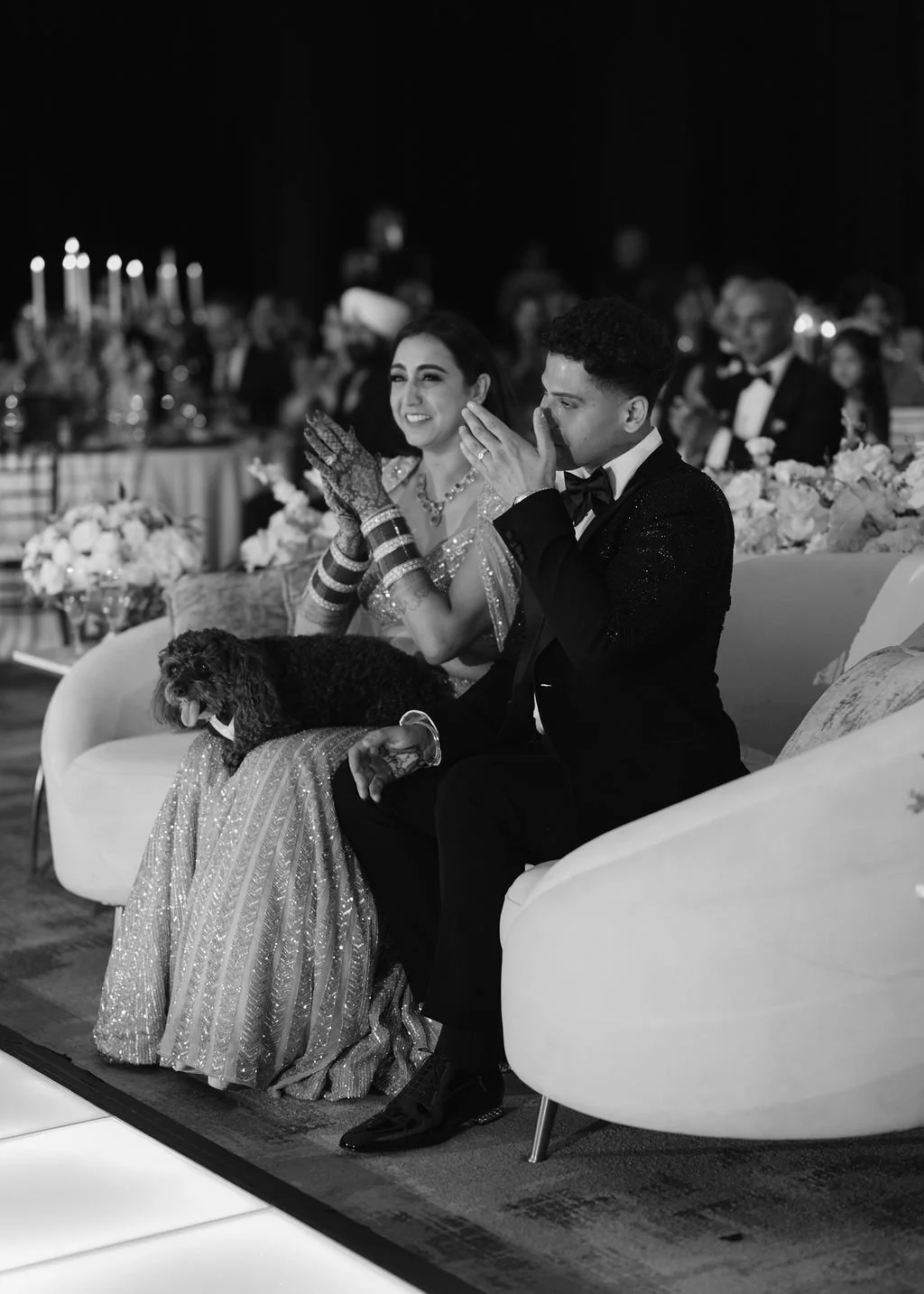 A black-and-white photo of a man and woman sitting on a white sofa at a formal event, possibly a wedding reception. The man in a tuxedo is whispering or speaking into the woman's ear. The woman is smiling, wearing traditional attire with jewelry and henna on her hands. They are seated next to a dog resting on the woman's lap. The background shows other guests seated at tables decorated with flowers and candles.