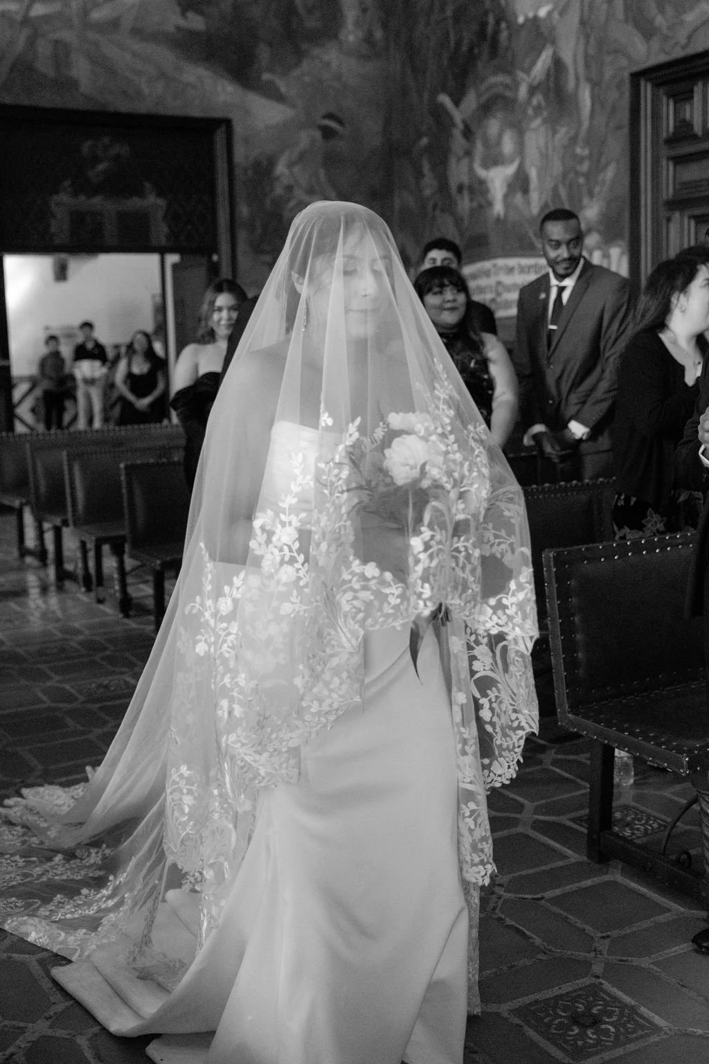 A bride wearing a veil and holding a flower bouquet during a wedding ceremony in a decorated church with seated guests in the background.