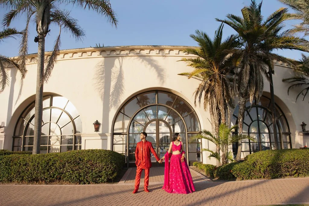 A couple in bright traditional Indian attire holding hands in front of a white building with large arched windows and palm trees.