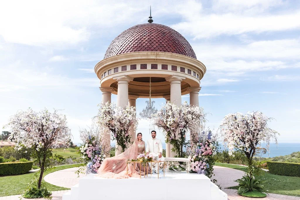 A wedding ceremony taking place outdoors under a domed pavilion with a red roof, surrounded by blooming cherry blossom trees, with a view of the ocean in the background.