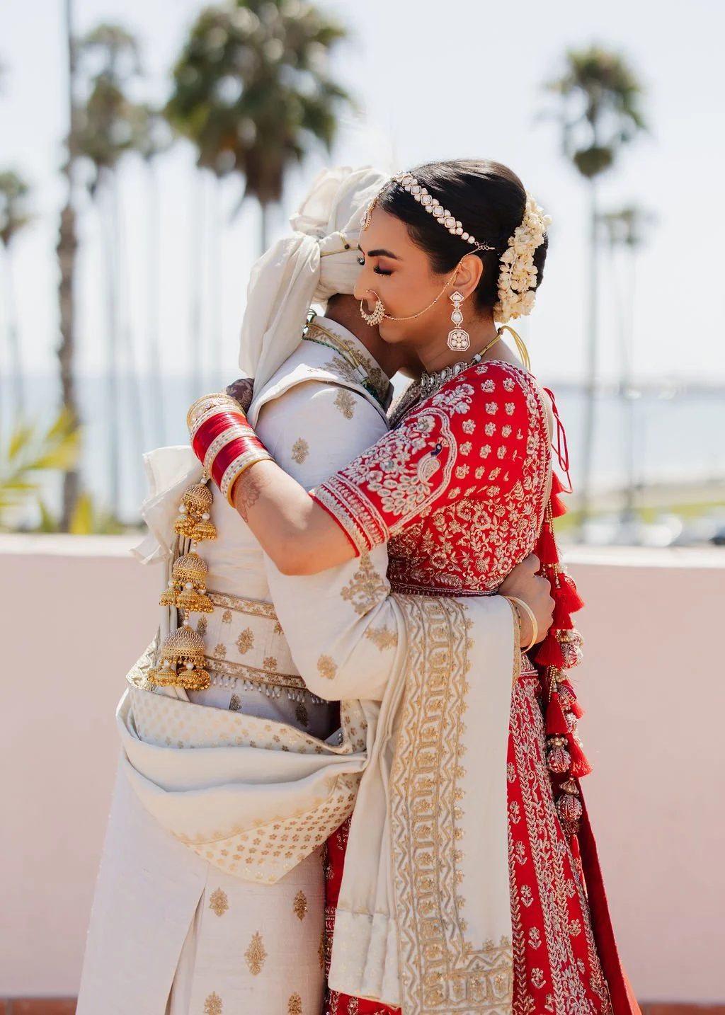 A bride and groom embrace outdoors, wearing traditional Indian wedding attire, with palm trees and a marina in the background.