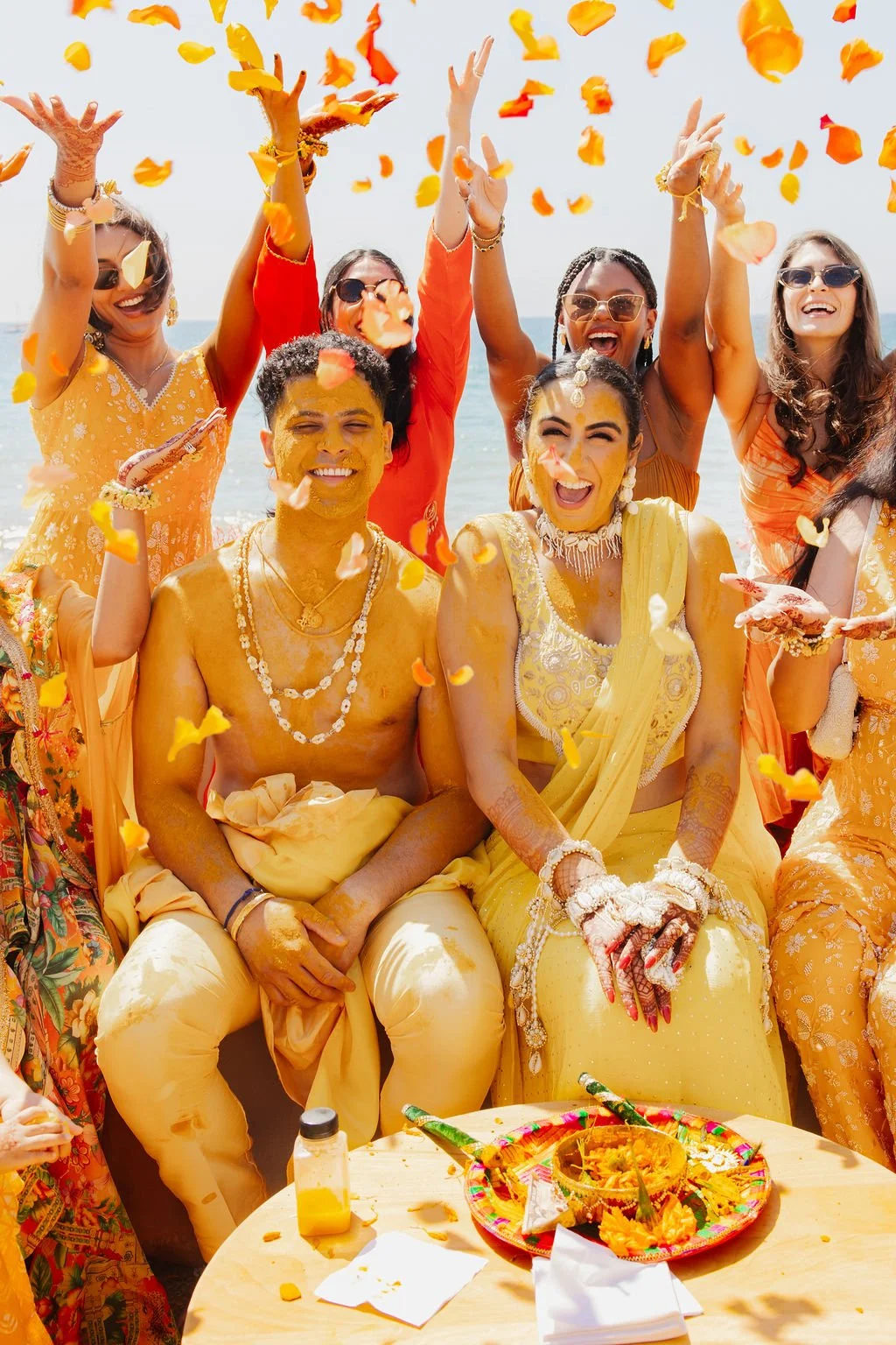 Group of people celebrating a traditional Indian ceremony by the beach, with yellow and orange floral clothing, throwing flower petals, and enjoying festive food.