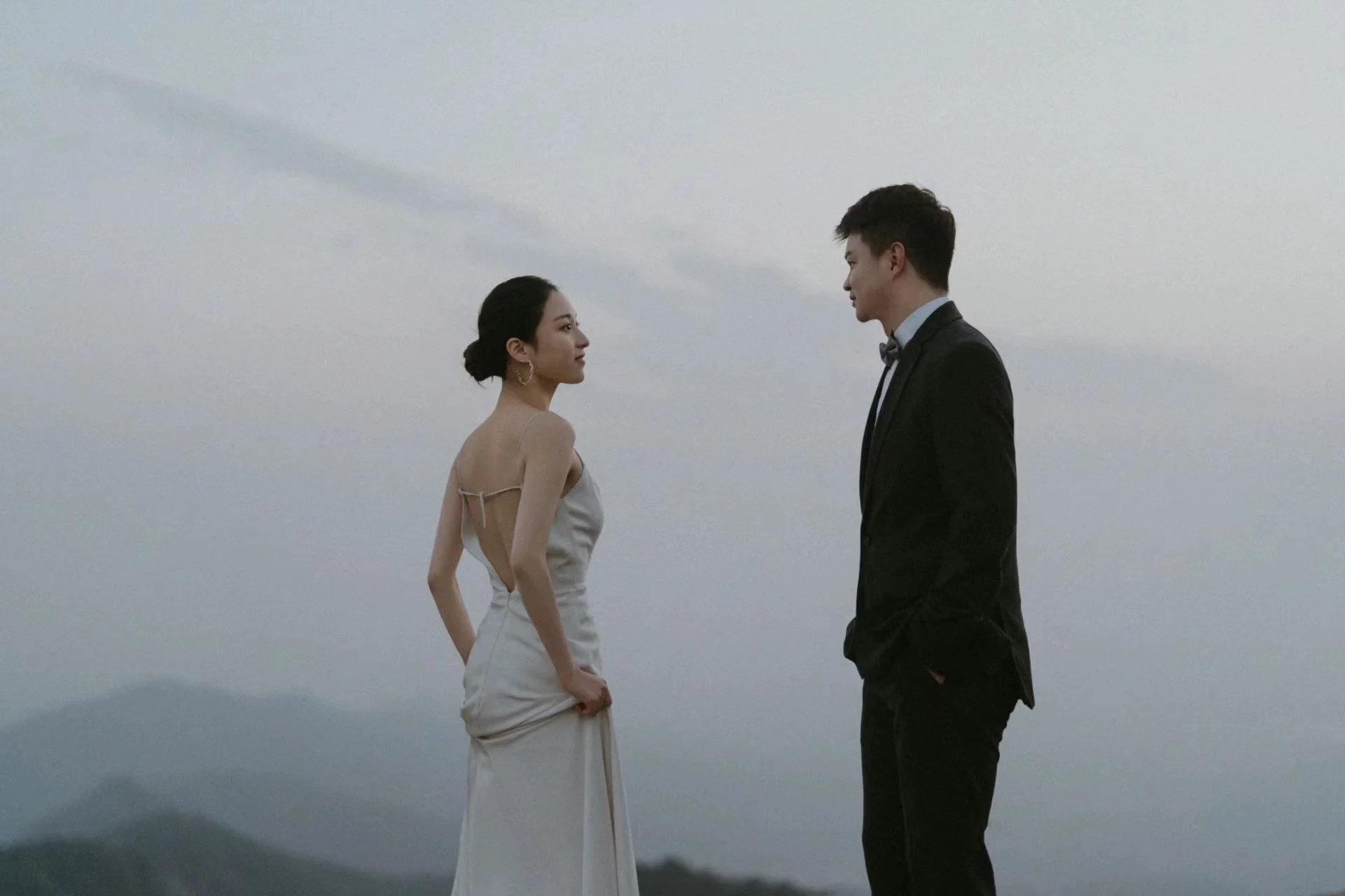 A woman in a white dress and a man in a black tuxedo facing each other outdoors during sunset or dusk.