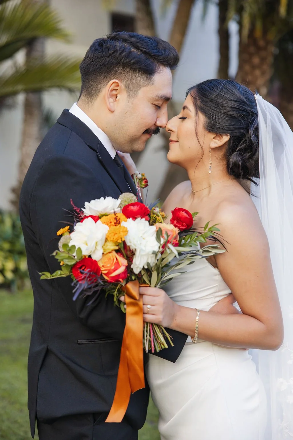 A newlywed couple stands close with their foreheads touching, smiling softly. The groom is wearing a black suit and tie, holding a colorful bouquet of flowers. The bride is in a strapless white wedding gown with a veil, wearing earrings and a bracele
