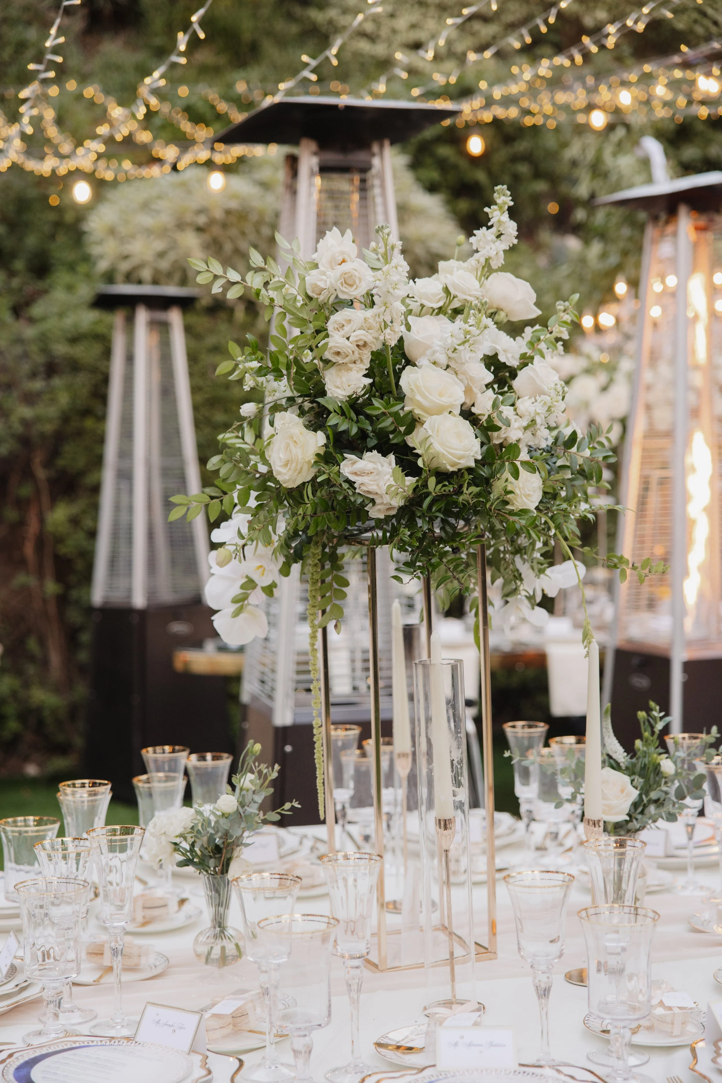 Elegant outdoor table setting with tall floral centerpiece of white roses and greenery, surrounded by glassware and candles, under string lights and patio heaters.