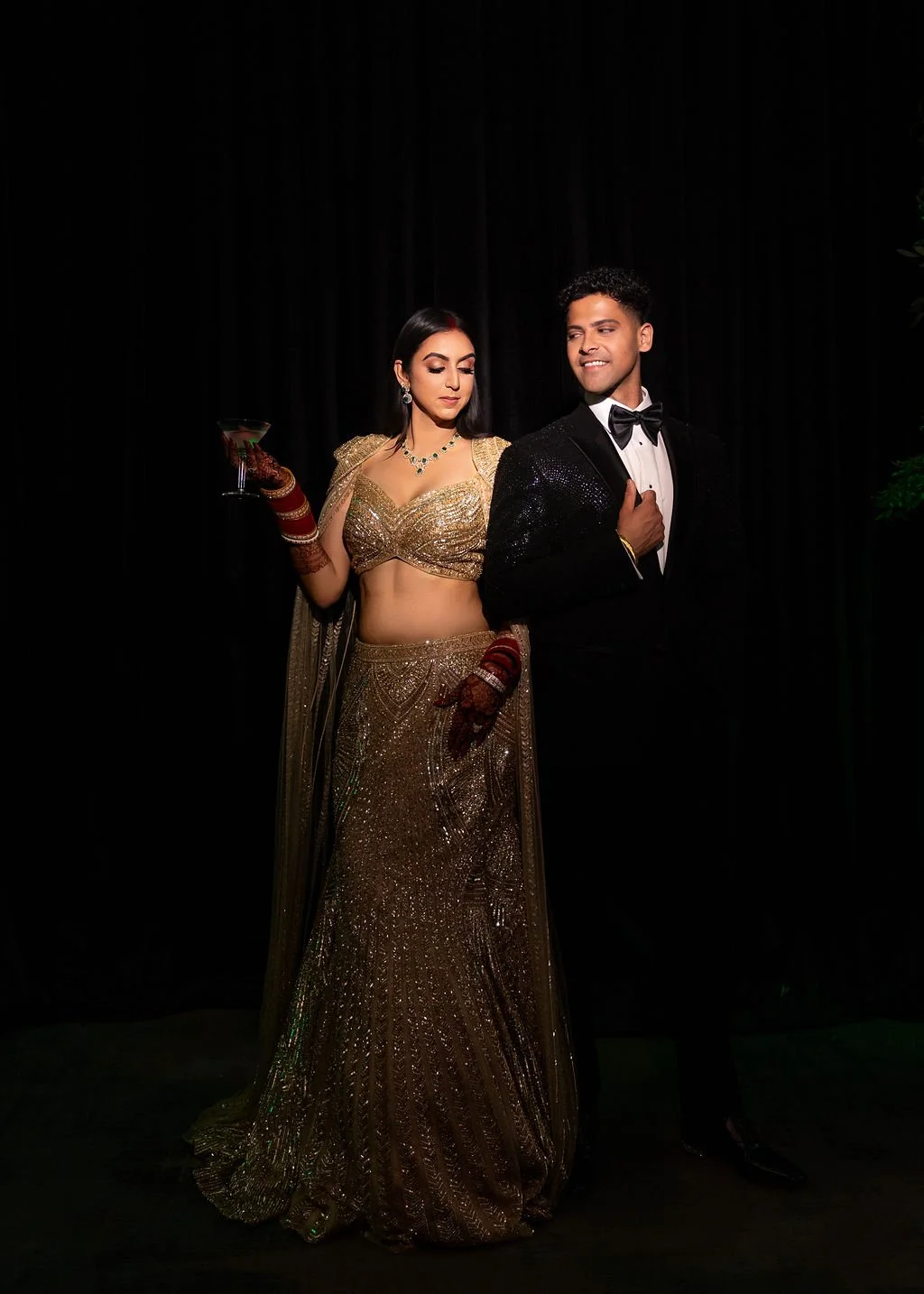 A couple dressed in elegant formal attire posing against a dark background, with the woman wearing a gold embellished saree and the man in a black tuxedo with a bow tie.