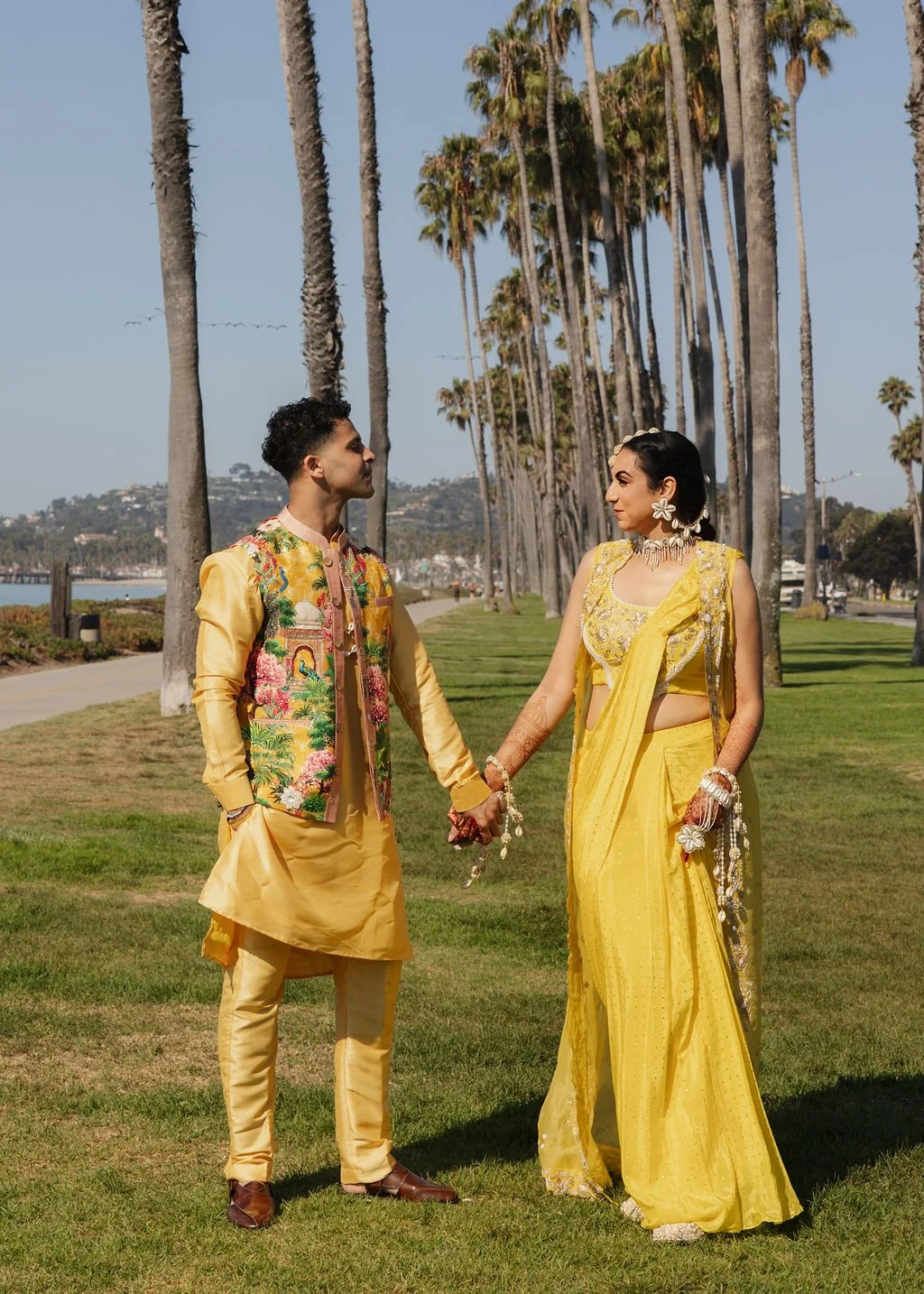 A couple dressed in traditional Indian attire holding hands on a grassy path lined with tall palm trees, with a waterfront and hills in the background.