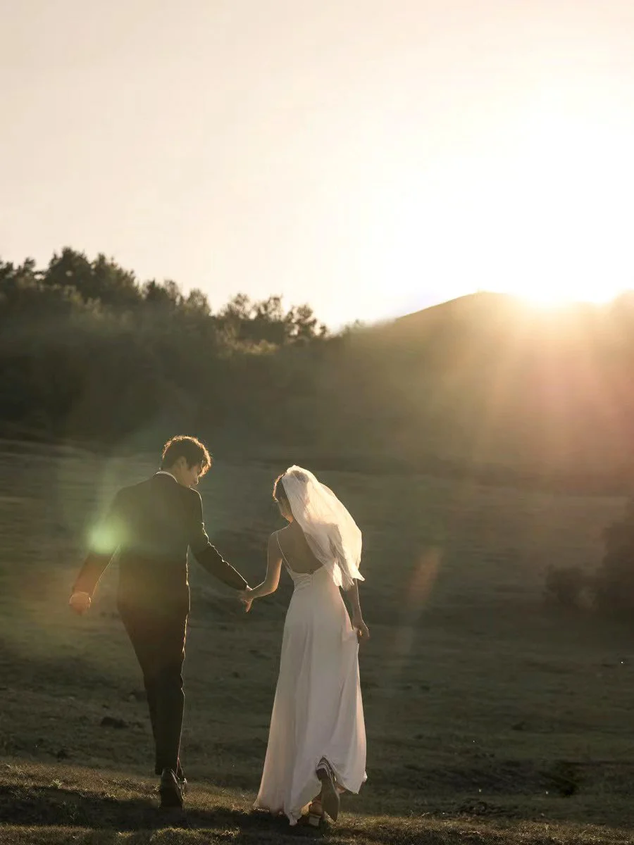 A couple dressed in wedding attire, holding hands and walking outdoors at sunset or sunrise in a scenic natural setting.