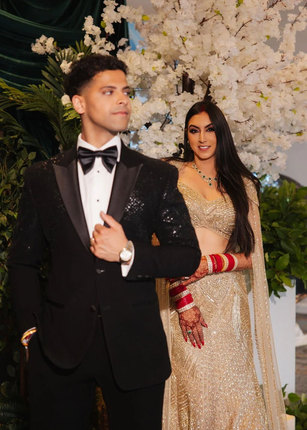 A man in a tuxedo with a bow tie walking in front of a woman dressed in a gold Indian saree, with white flowers and greenery in the background.