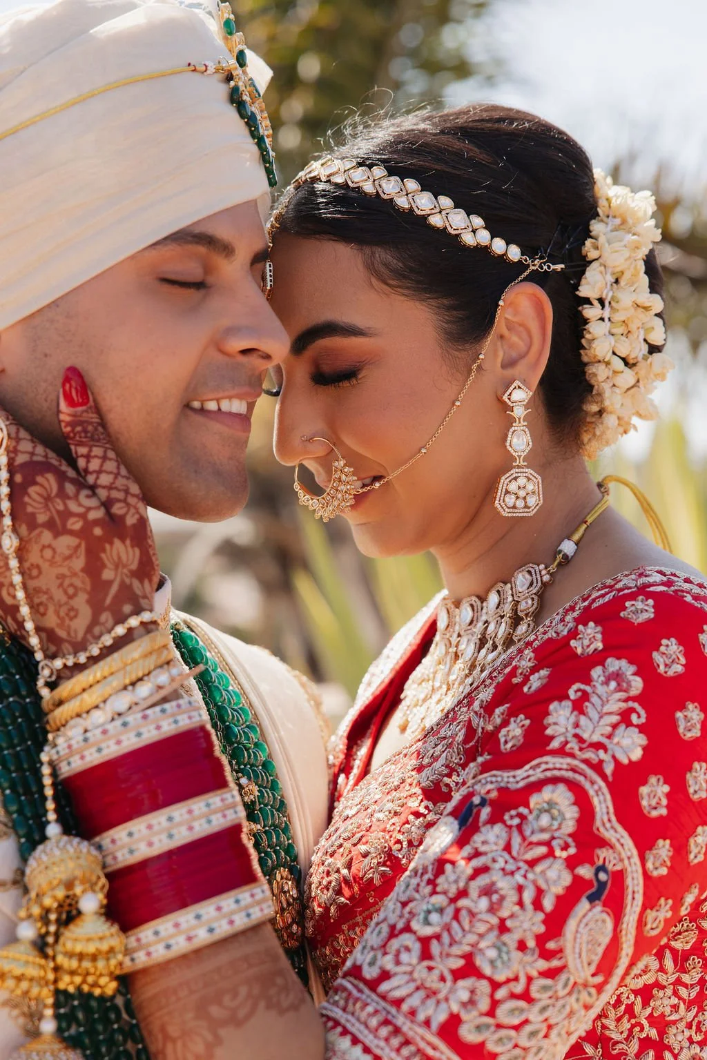 A couple in traditional Indian wedding attire sharing an affectionate moment with foreheads touching, amidst natural outdoor surroundings.