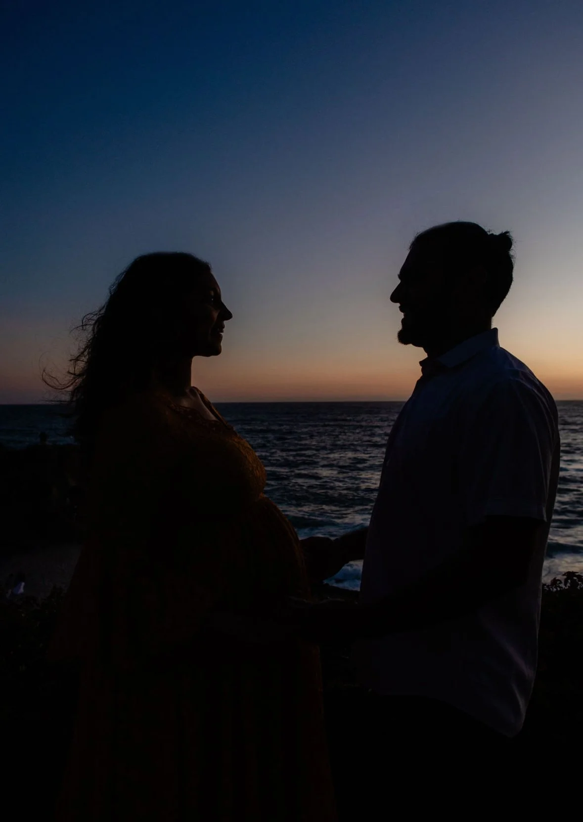 Silhouette of a couple holding hands by the ocean at sunset, facing each other with a colorful sky in the background.