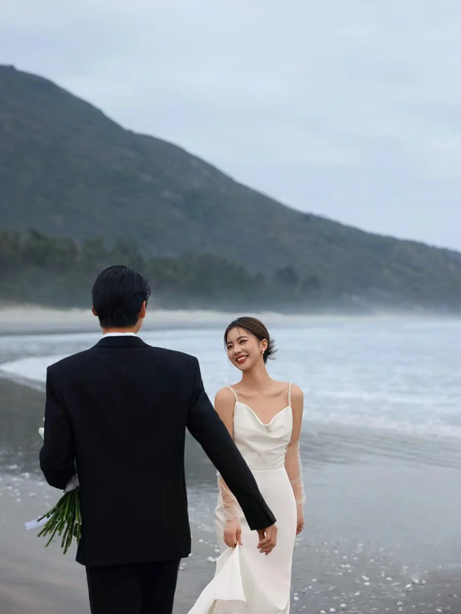 A bride and groom standing near the ocean on a beach, with mountains in the background. The bride is smiling, wearing a white wedding dress, and holding a part of her dress. The groom is dressed in a dark suit, holding a bouquet of flowers behind his