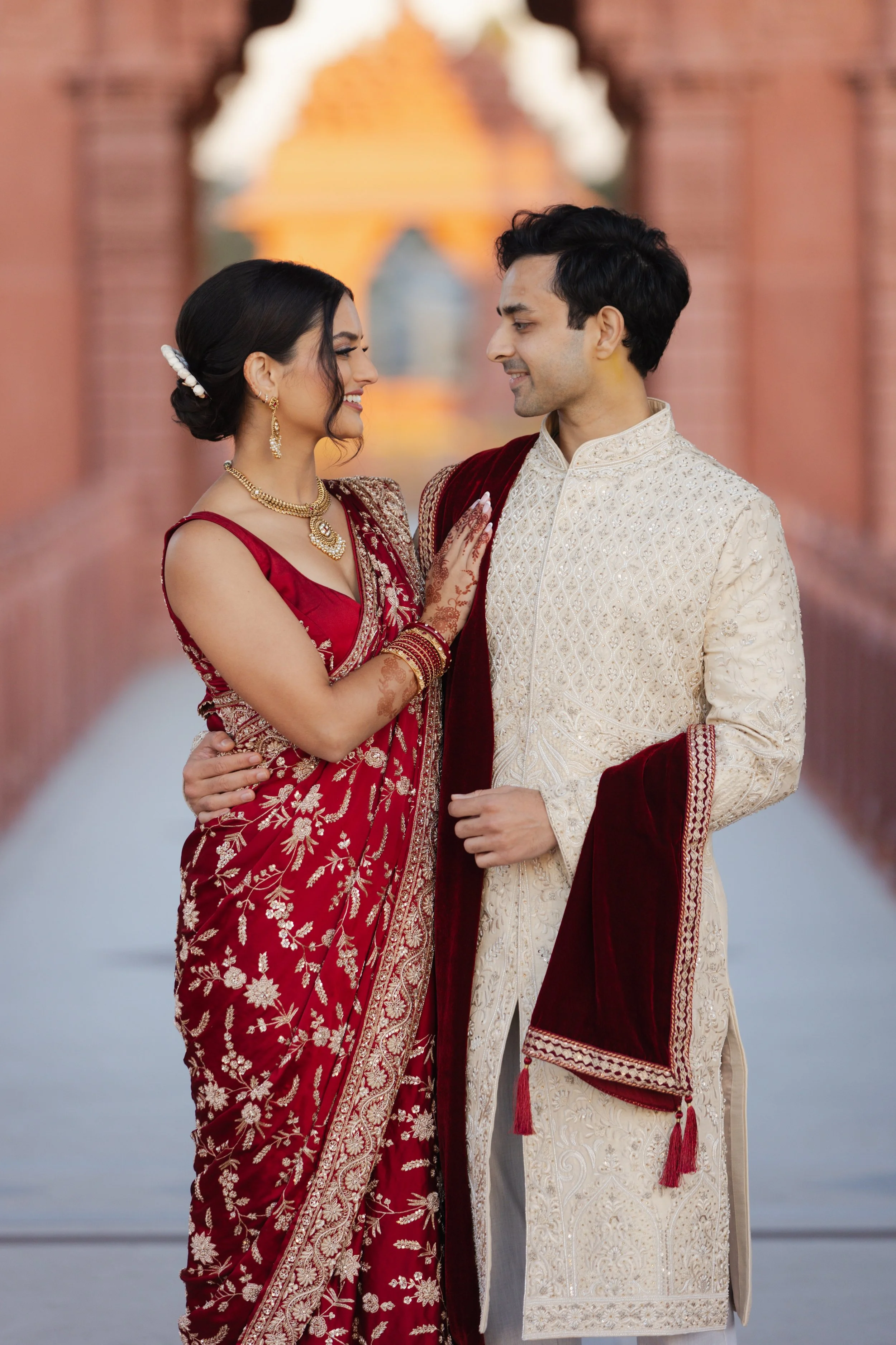 A couple in traditional Indian wedding attire, smiling at each other with the bride's hand on the groom's chest, standing on a bridge with an architectural background at BAPS in chino hills, california.