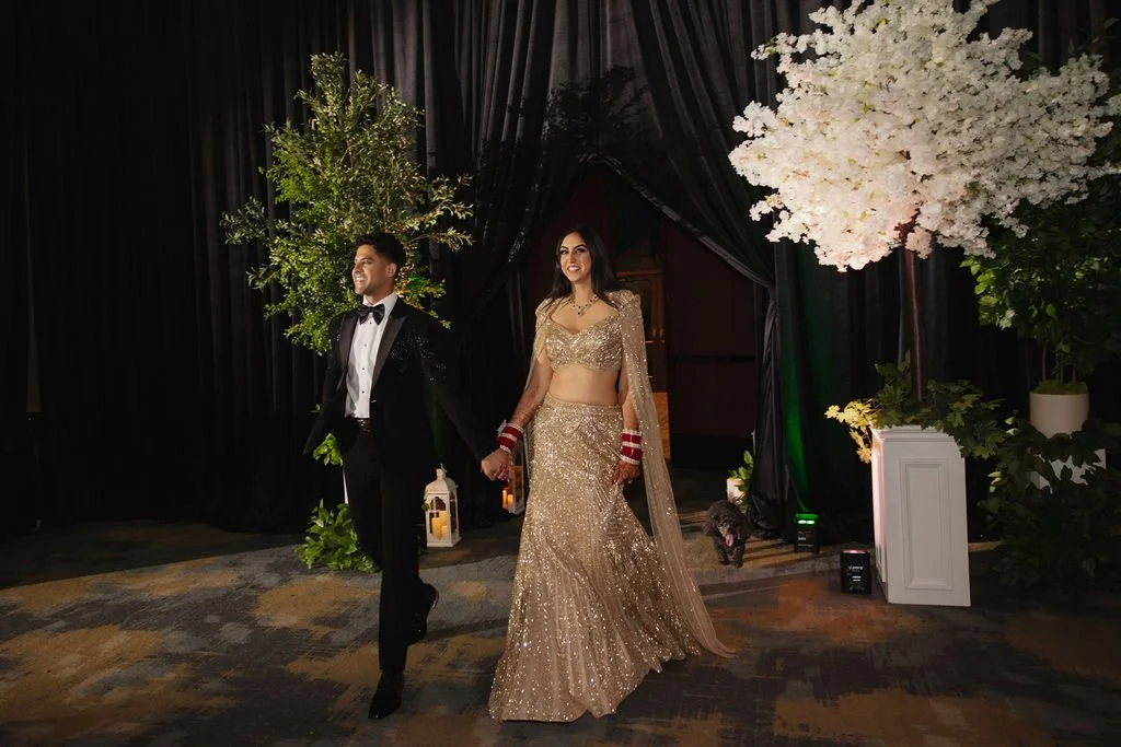 A man and woman holding hands while walking at a decorated event. The man is in a black tuxedo and the woman is in a gold Indian bridal outfit. There are floral arrangements and decorative lanterns around them.