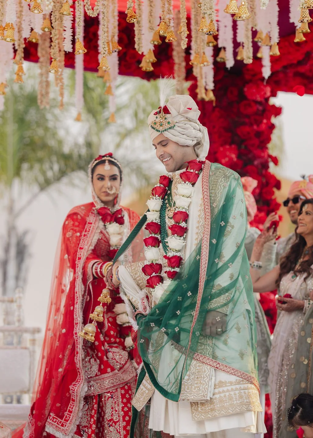 A groom dressed in traditional Indian wedding attire, including a turban and floral garland, smiles at a wedding ceremony under a decorated canopy. The bride is dressed in red with intricate embroidery, jewelry, and a veil, holding a sword. Several g