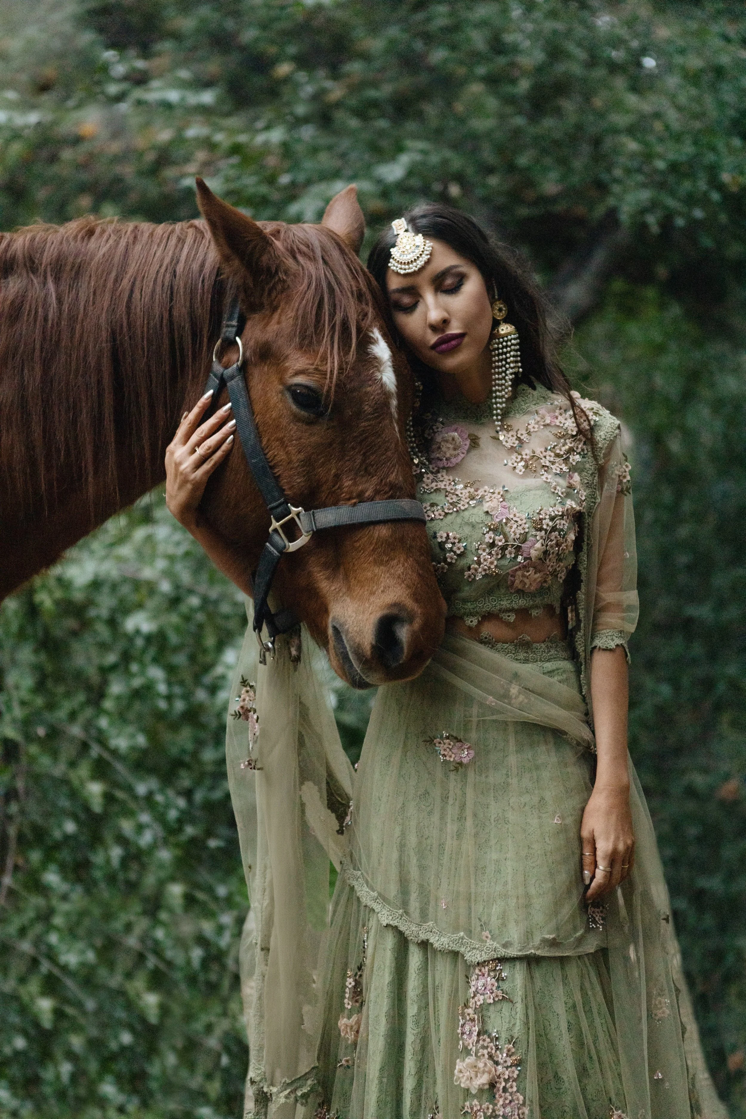 A woman dressed in an embroidered, sheer, pastel-green traditional outfit with floral designs, gently touching a brown horse's face outdoors.