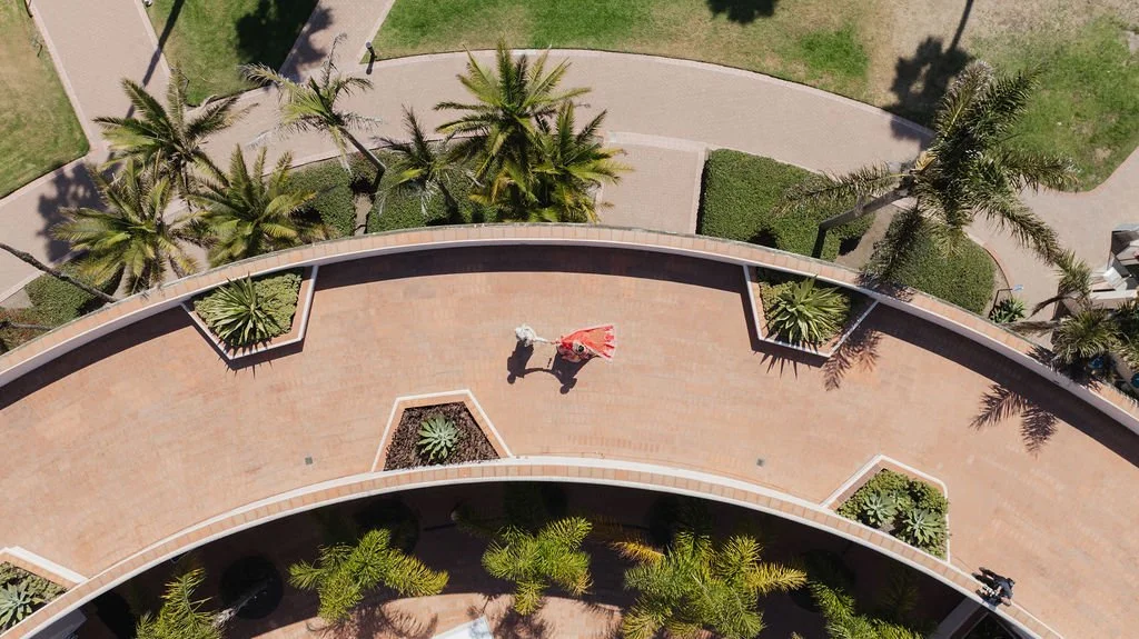 Aerial drone view of a first look where curved brick walkway with a person walking and holding a red umbrella, surrounded by palm trees and landscaped greenery.