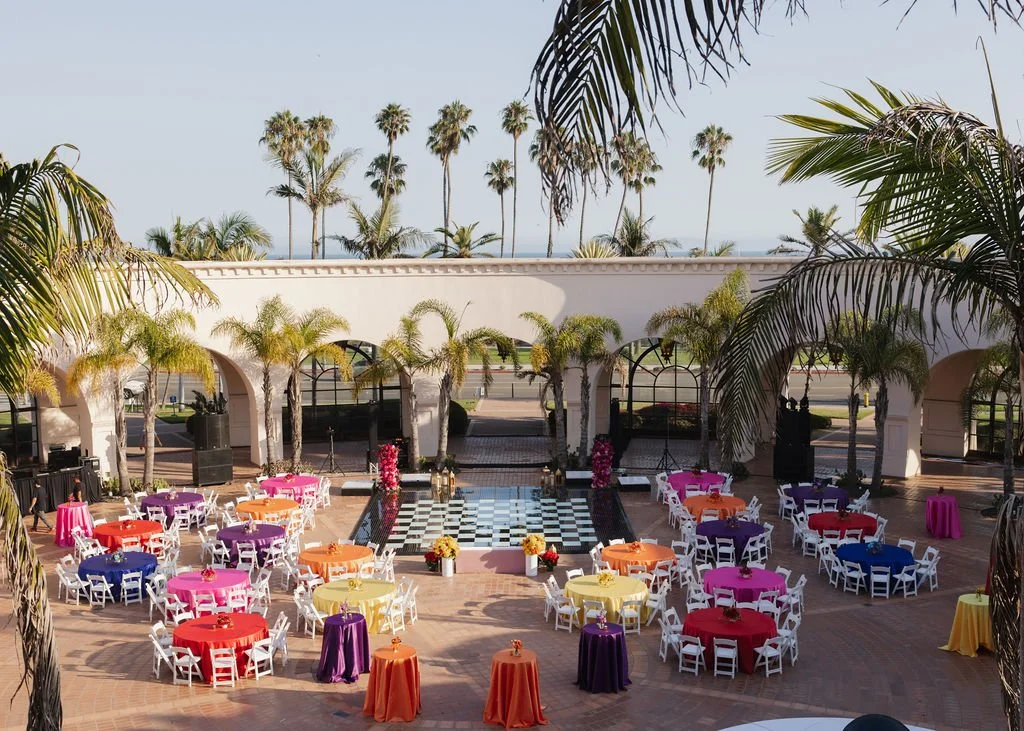 Outdoor event space with colorful round tables and white chairs, decorated with flowers. Palm trees surround the area, with a building and more palm trees in the background.
