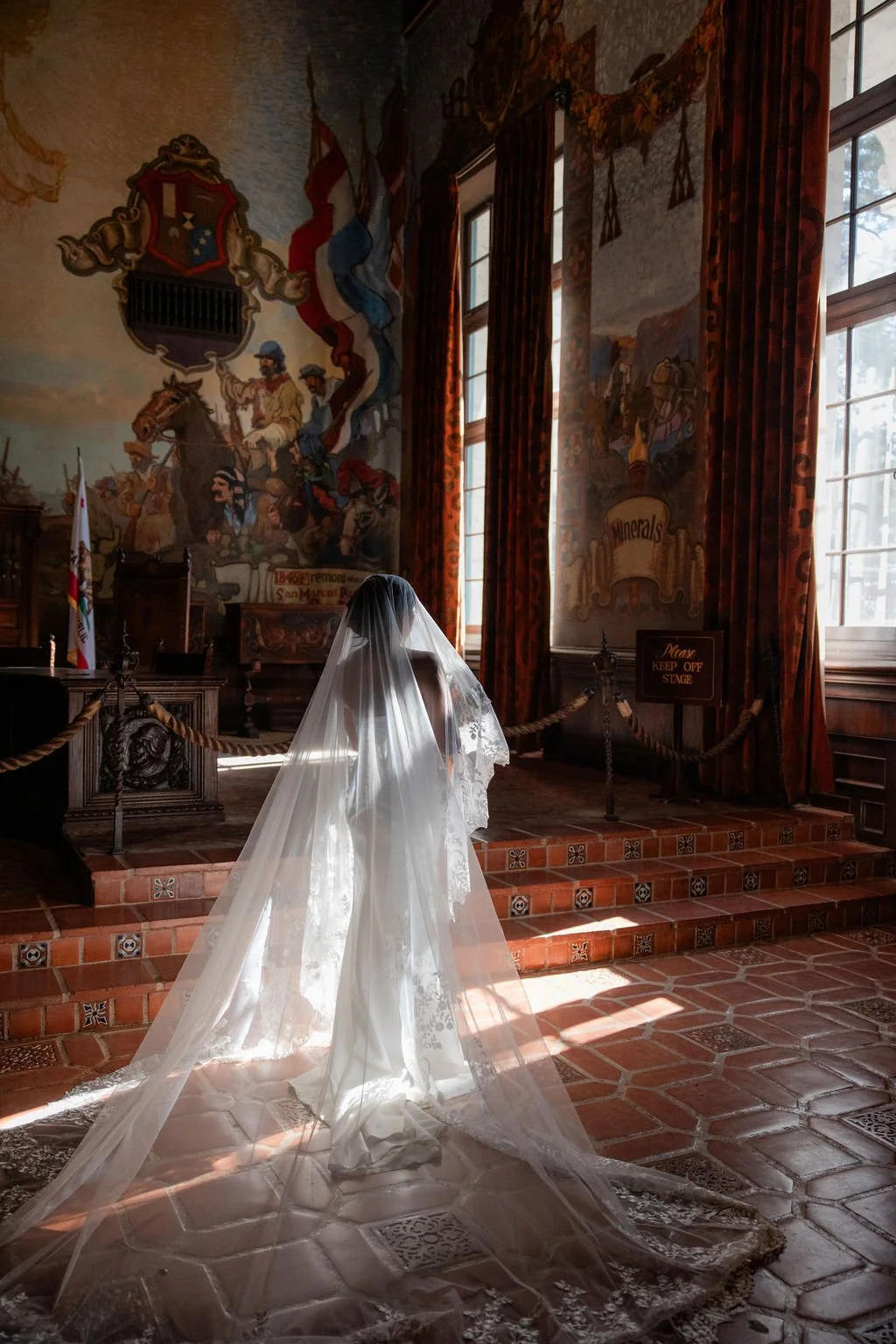 A bride in a white wedding dress and veil stands in an ornate historical room with large windows, colorful murals of circus performers on the walls, and a stage with a sign that says 'Please Keep Off Stage.' The room has high ceilings, hardwood floor