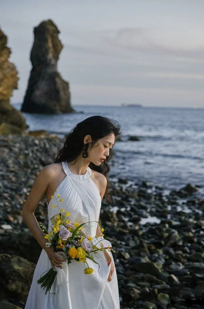 A woman in a white dress holding a bouquet of yellow and purple flowers on a rocky beach with sea stacks in the background.