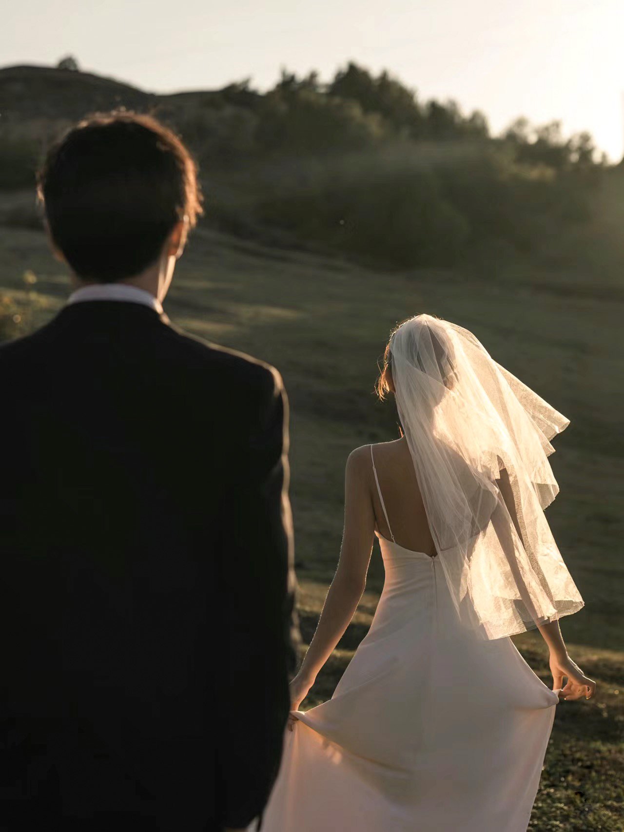A bride in a white wedding dress and veil standing outdoors with a groom in a black suit, on a grassy field with hills in the background, during sunset.