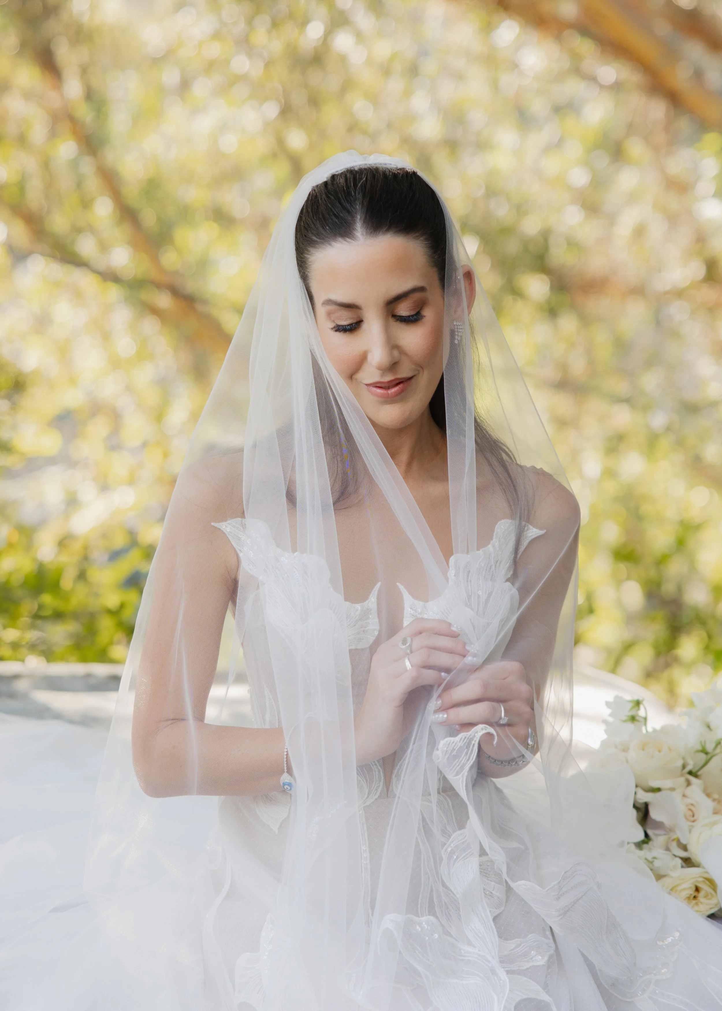 Bride with long dark hair in a white wedding dress and veil, standing outdoors with blurry trees and sunlight in the background.