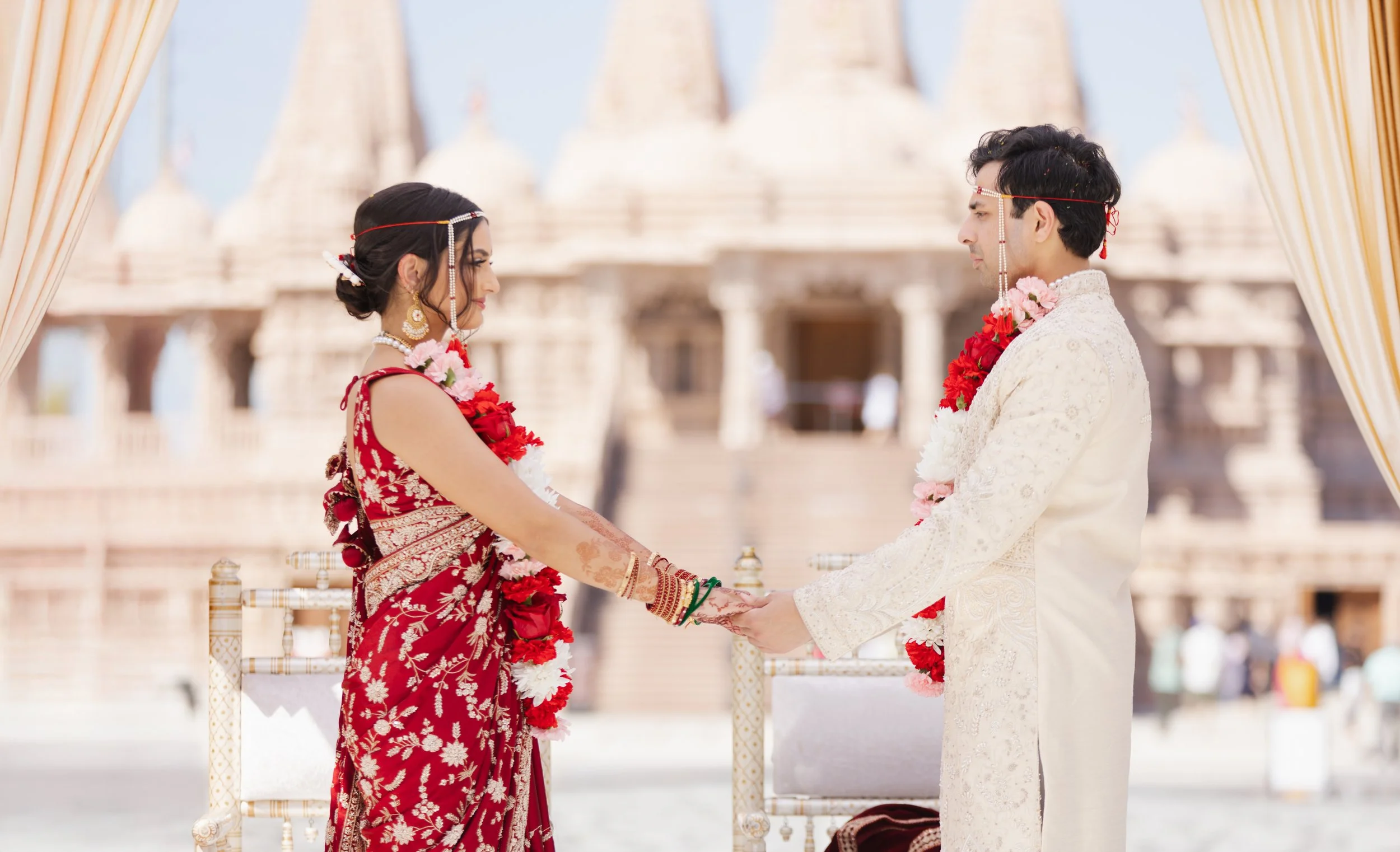 Indian bride and groom holding hands during a wedding ceremony in front of a temple, dressed in traditional attire, with flowers and wedding accessories at BAPS in chino hills, california.