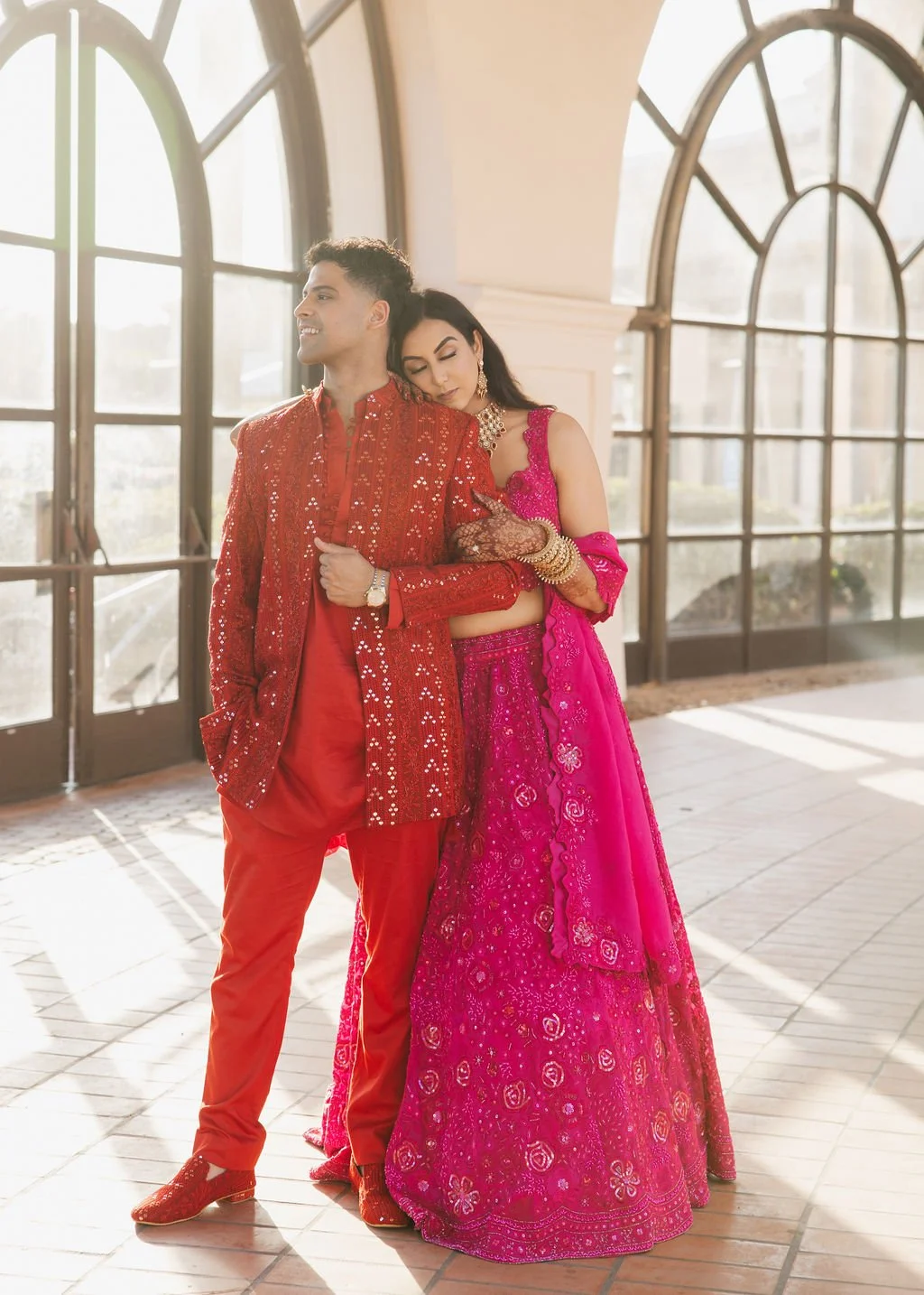A couple in colorful traditional Indian attire standing in front of large glass windows before their sangeet, with sunlight illuminating them. The bride leans her head on the groom's shoulder, and they both appear happy and affectionate.