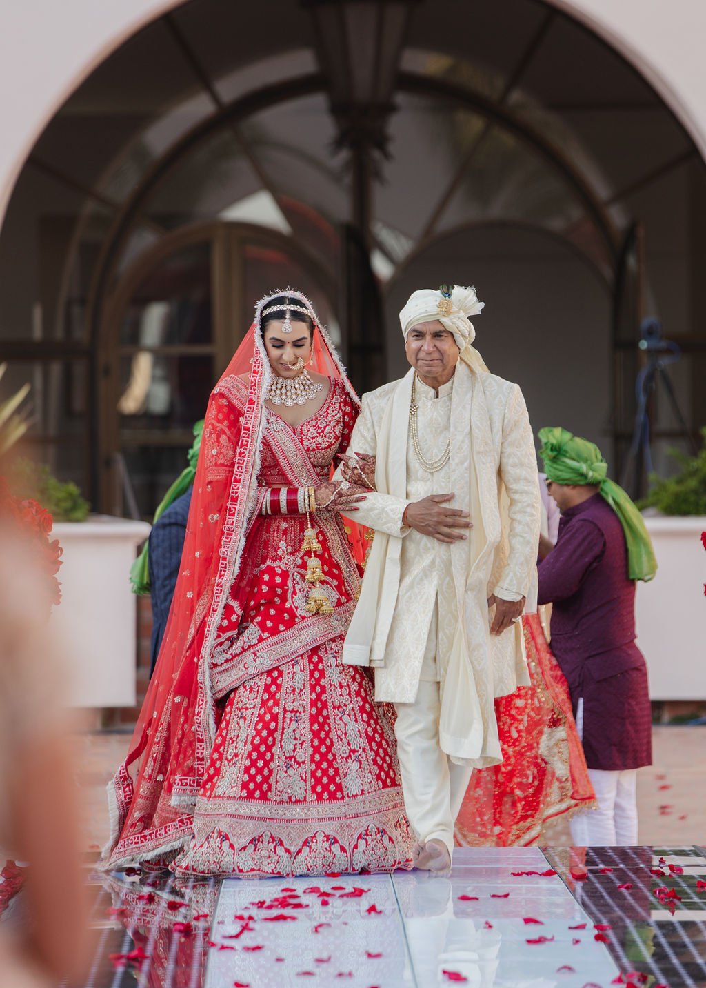 A bride in a red traditional wedding dress and jewelry walking with the aisle at her hindu ceremony wedding with her dad/father wearing an off-white sherwani and turban, during a wedding ceremony. Rose petals are scattered on the floor.