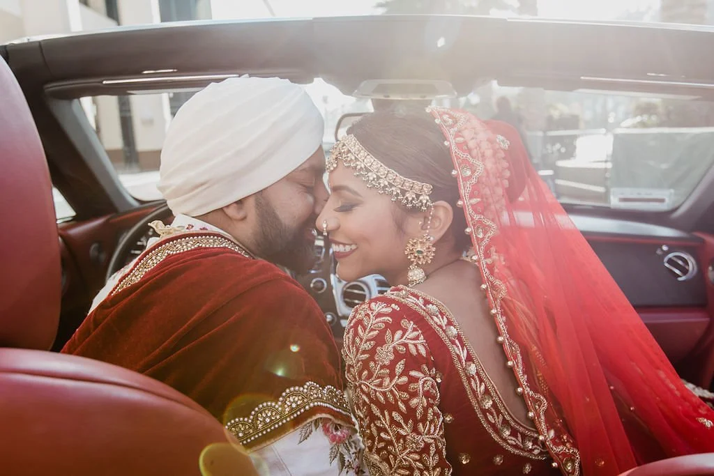 A newlywed couple in traditional wedding attire sitting in a car, smiling and touching foreheads, with sunlight streaming in.