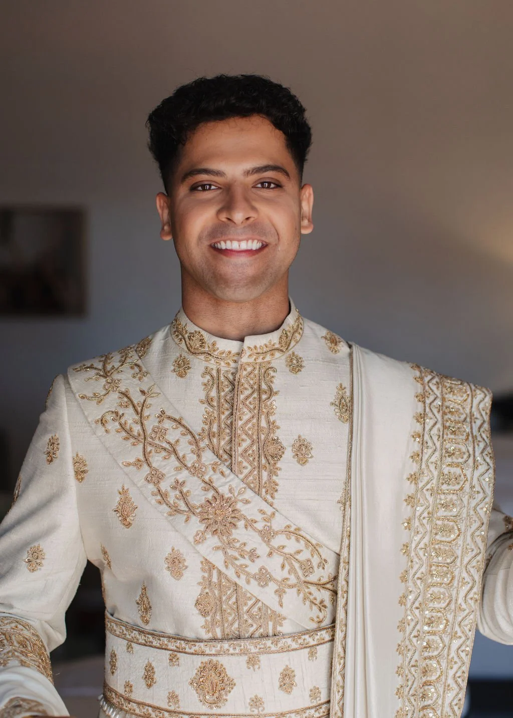 A groom wearing an intricately embroidered cream-colored traditional Indian outfit, smiling with a neutral indoor background at Hilton Santa Barbara beachfront in california.