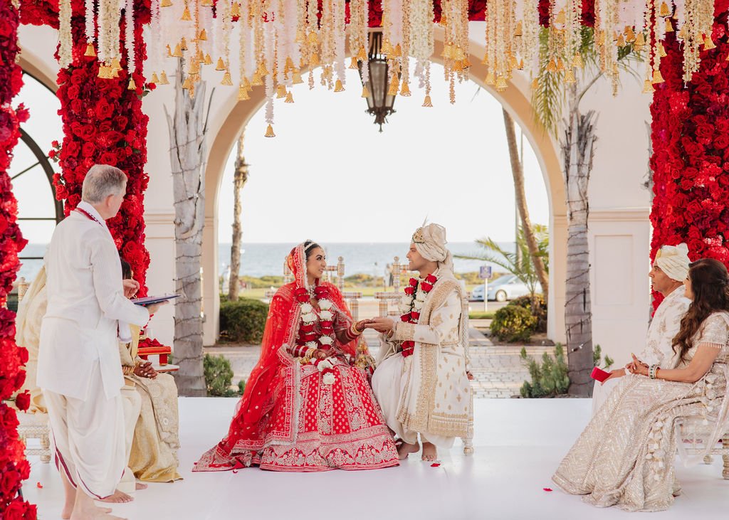 Luxury high-end Indian wedding ceremony with bride and groom exchanging garlands under floral arch, surrounded by family, on a sunny outdoor setting near a beach in Hilton Santa Barbara.