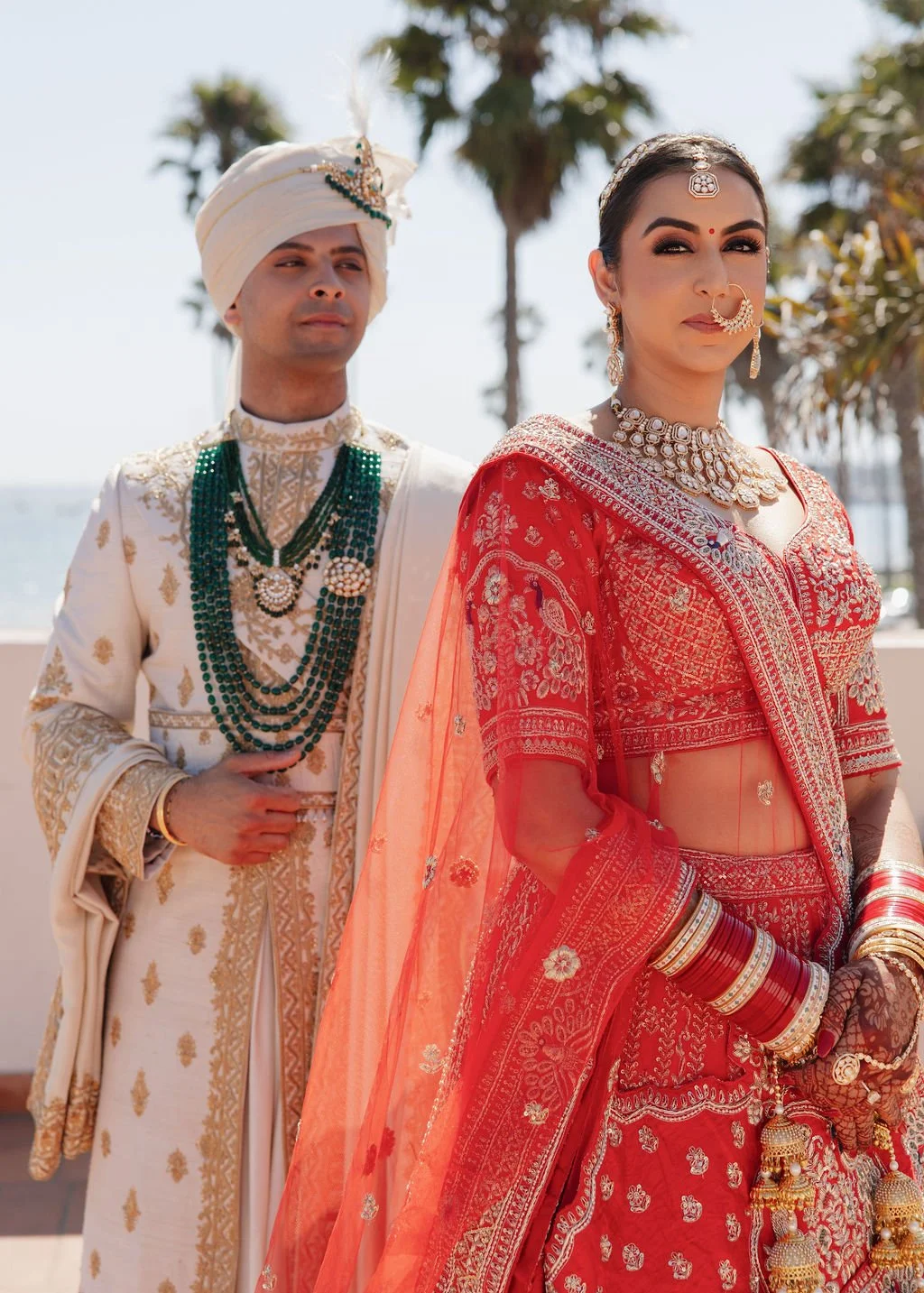 A couple in traditional Indian wedding attire after their first look , with the woman in a red lehenga and the man in a cream-colored sherwani and turban, standing outdoors with trees and water in the background in Hilton Santa Barbara beachfront.