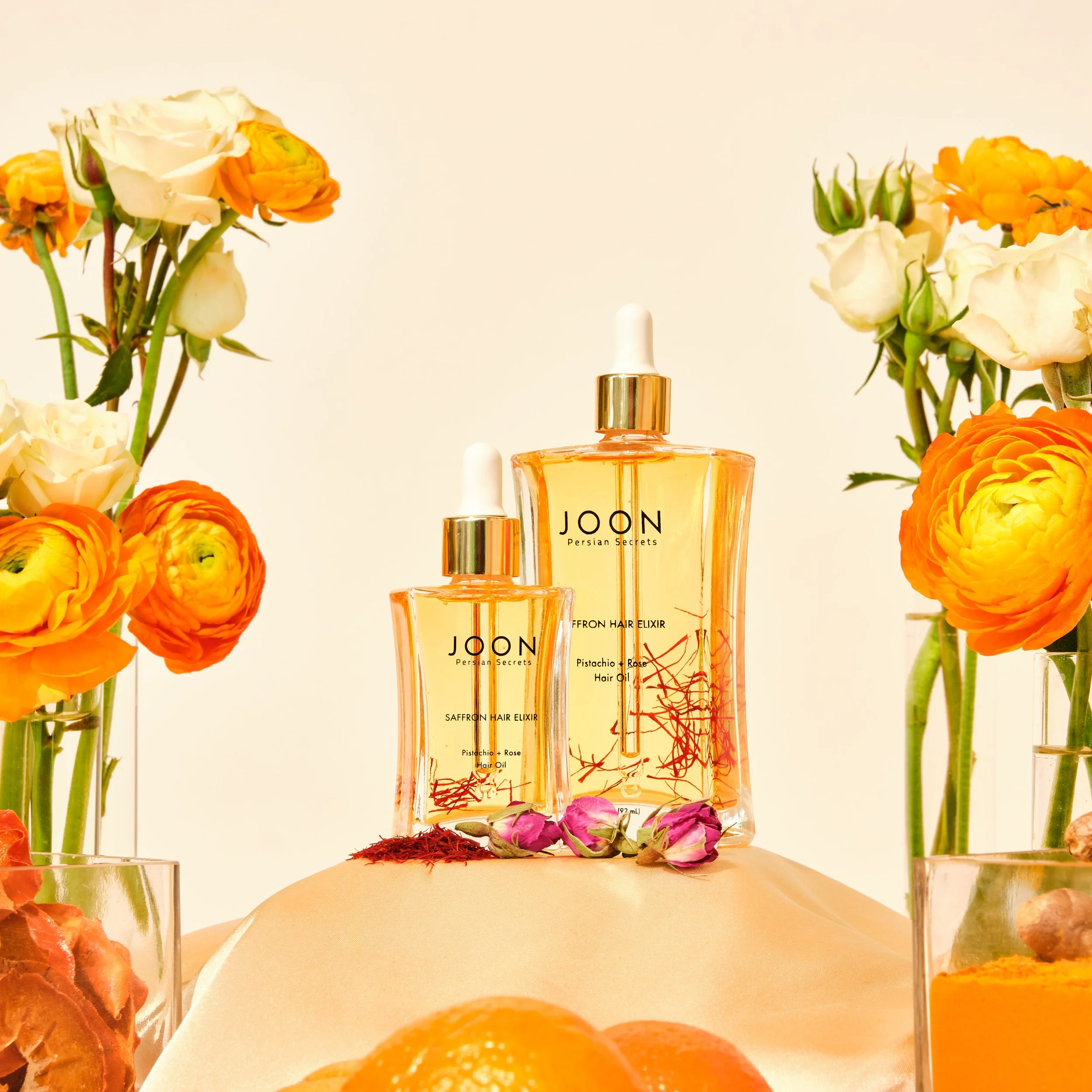 Two bottles of JOON Persian Secrets hair elixir, one larger and one smaller, surrounded by orange and white flowers in glass vases and pieces of fruit, set on a beige surface against a plain background.